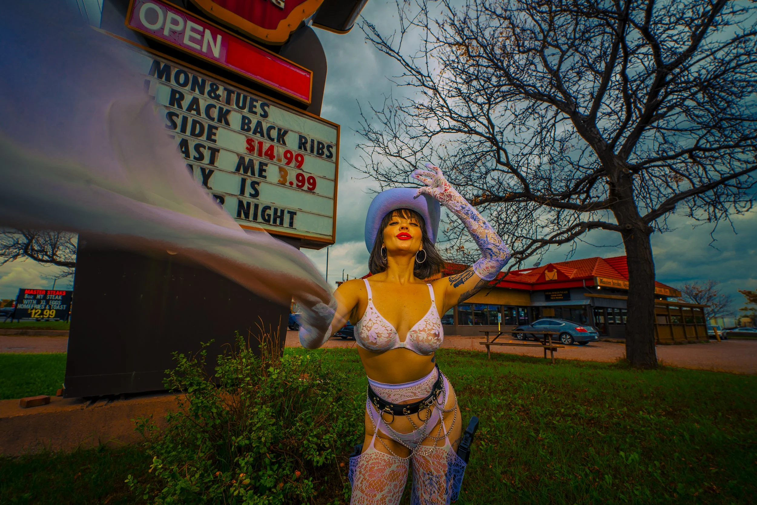 A woman dressed in revealing lingerie with lace bohemian style and tattoos, wearing a purple cowboy hat and large hoop earrings, poses with her hand on her hat in front of a fast food restaurant and a signboard with prices, with a bare-branched tree 