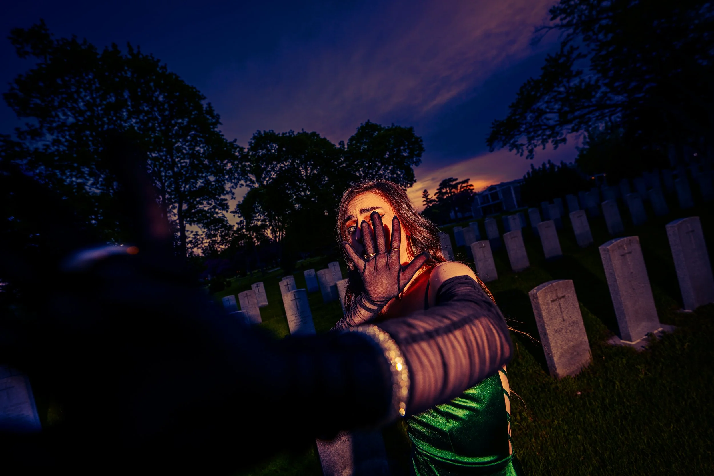A person taking a selfie in a cemetery at dusk, with gravestones and trees in the background.