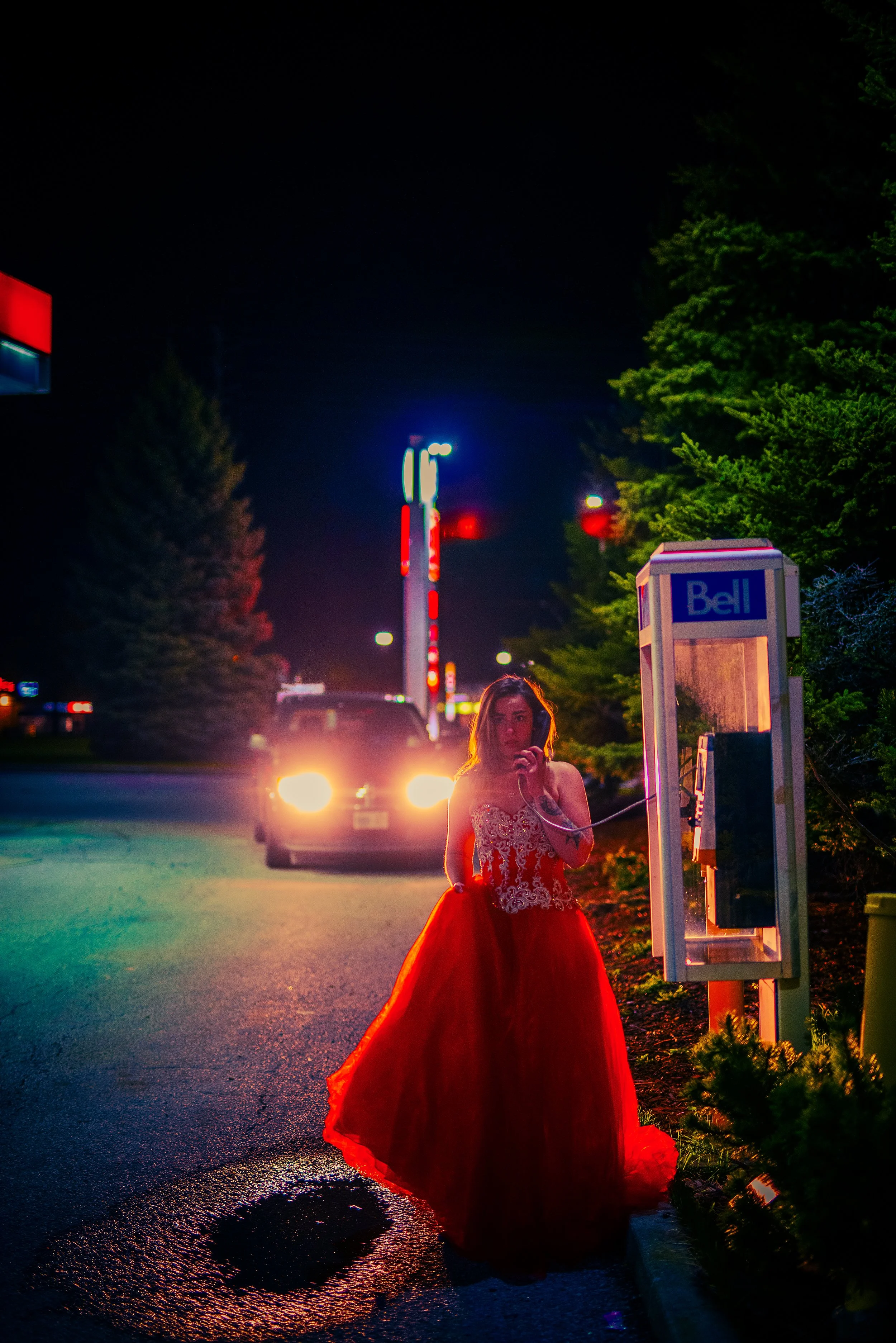 A woman wearing a red ball gown and lace top standing at night by a phone booth with a bell logo, under colorful streetlights, with a car approaching from behind with headlights on.