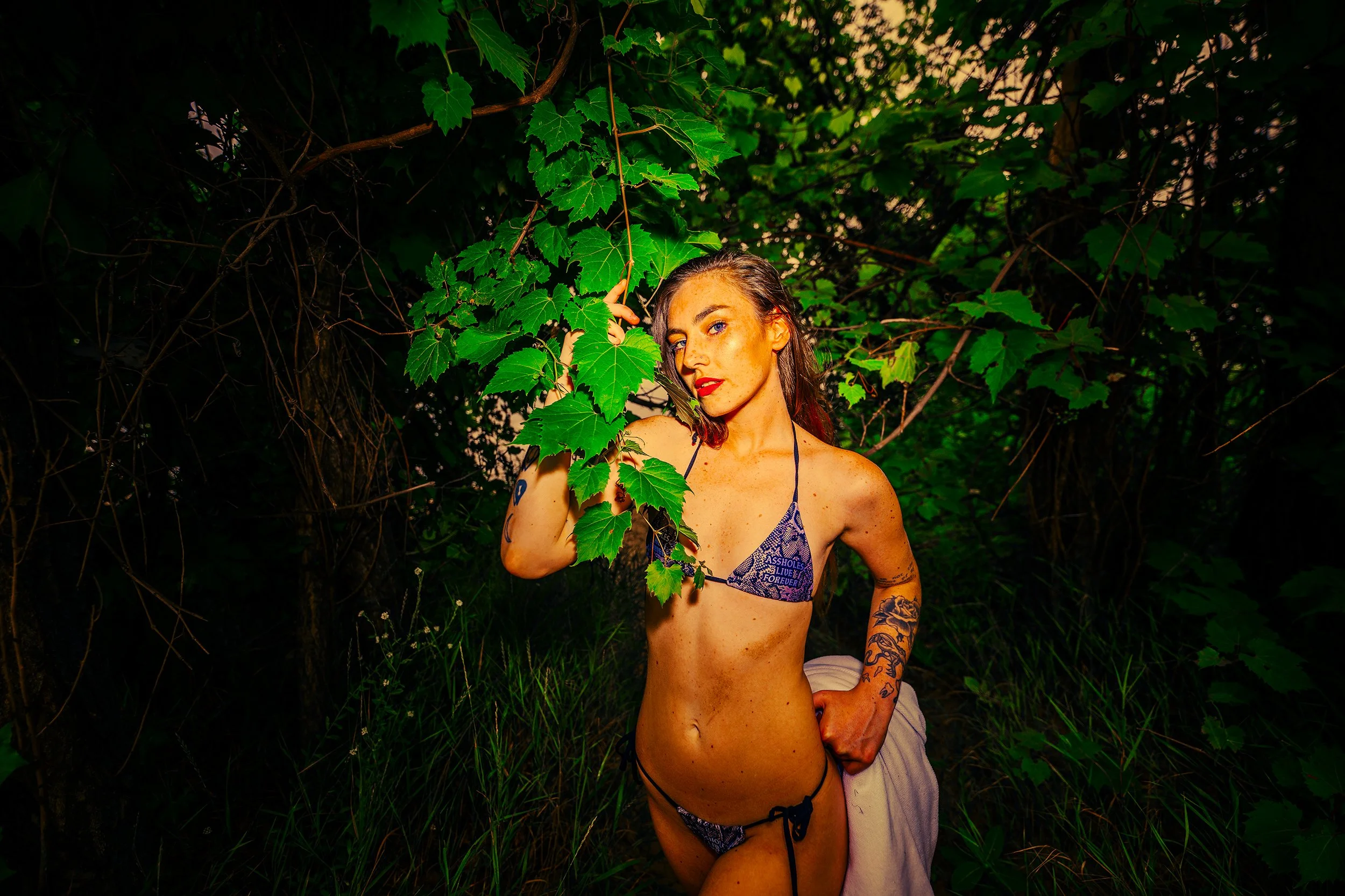 A woman with tattoos, wearing lingerie, is kneeling outdoors among green foliage, holding onto a vine, with a partial view of her face and body visible.