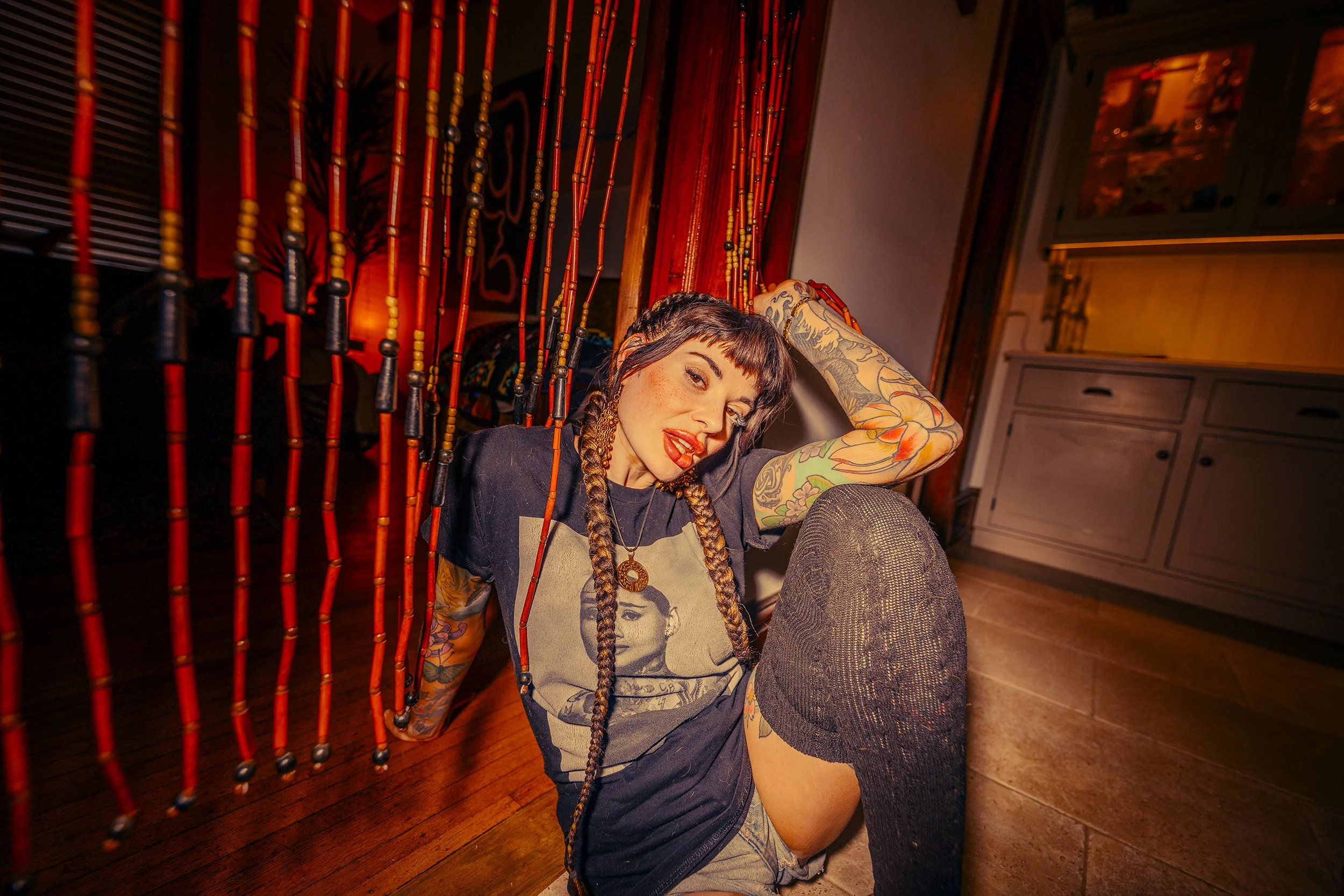A woman with tattoos and braids sitting on the floor, smiling at the camera, indoors with red bead curtains behind her and a painting on the wall.