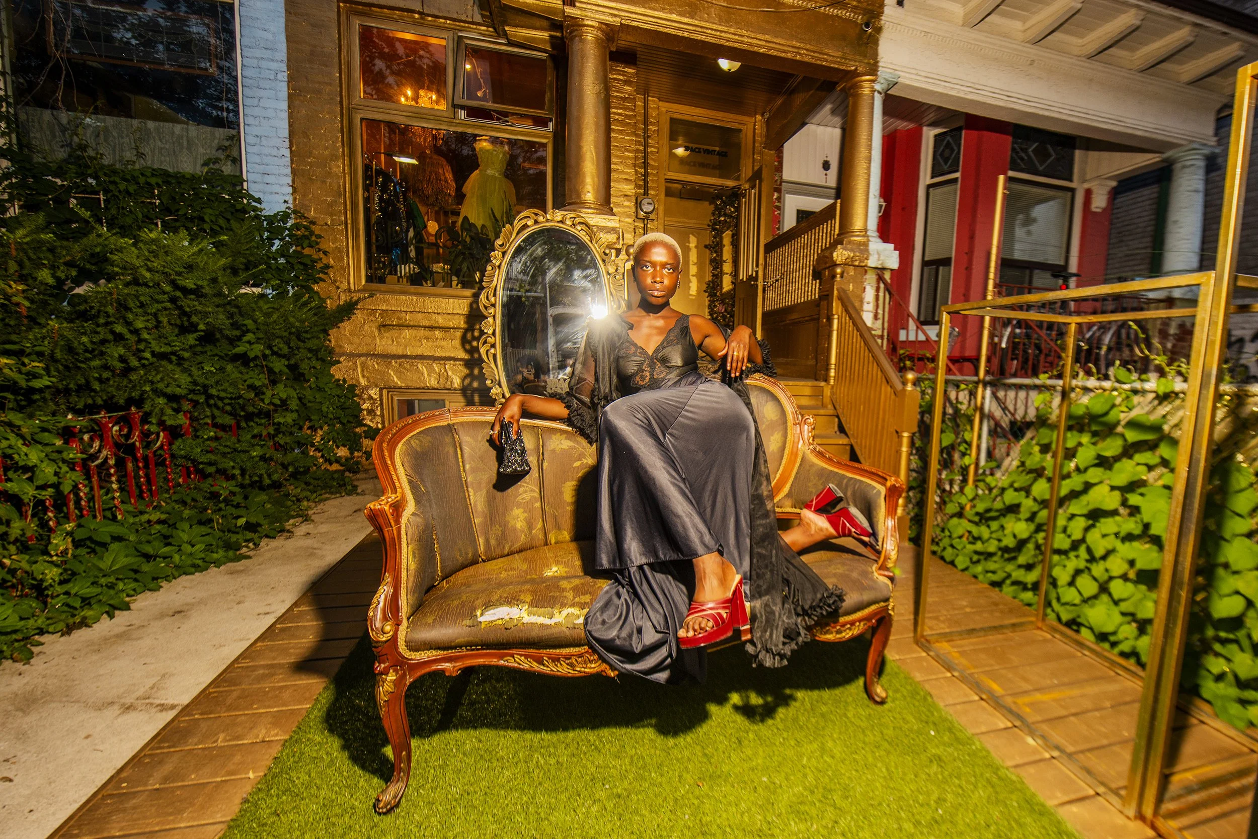 A woman with platinum blonde hair sitting on a vintage sofa outside a house at night, wearing a black dress and red heels, holding a small handbag, with a large mirror and various decorative elements behind her.