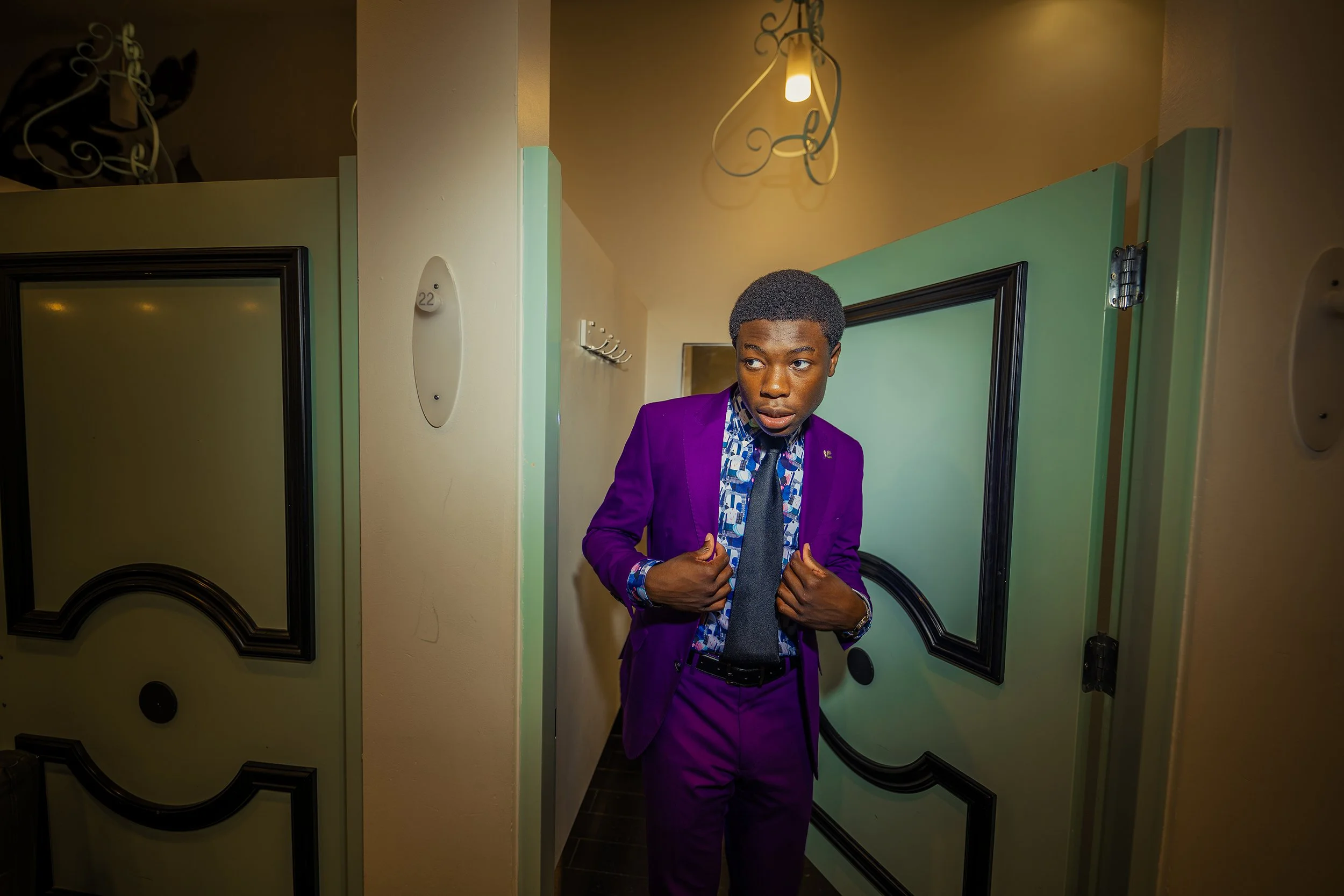 A young man in a purple suit with a patterned shirt and a black tie standing in a room with colored walls and decorative lighting. He is holding his jacket lapels and looking to his left.