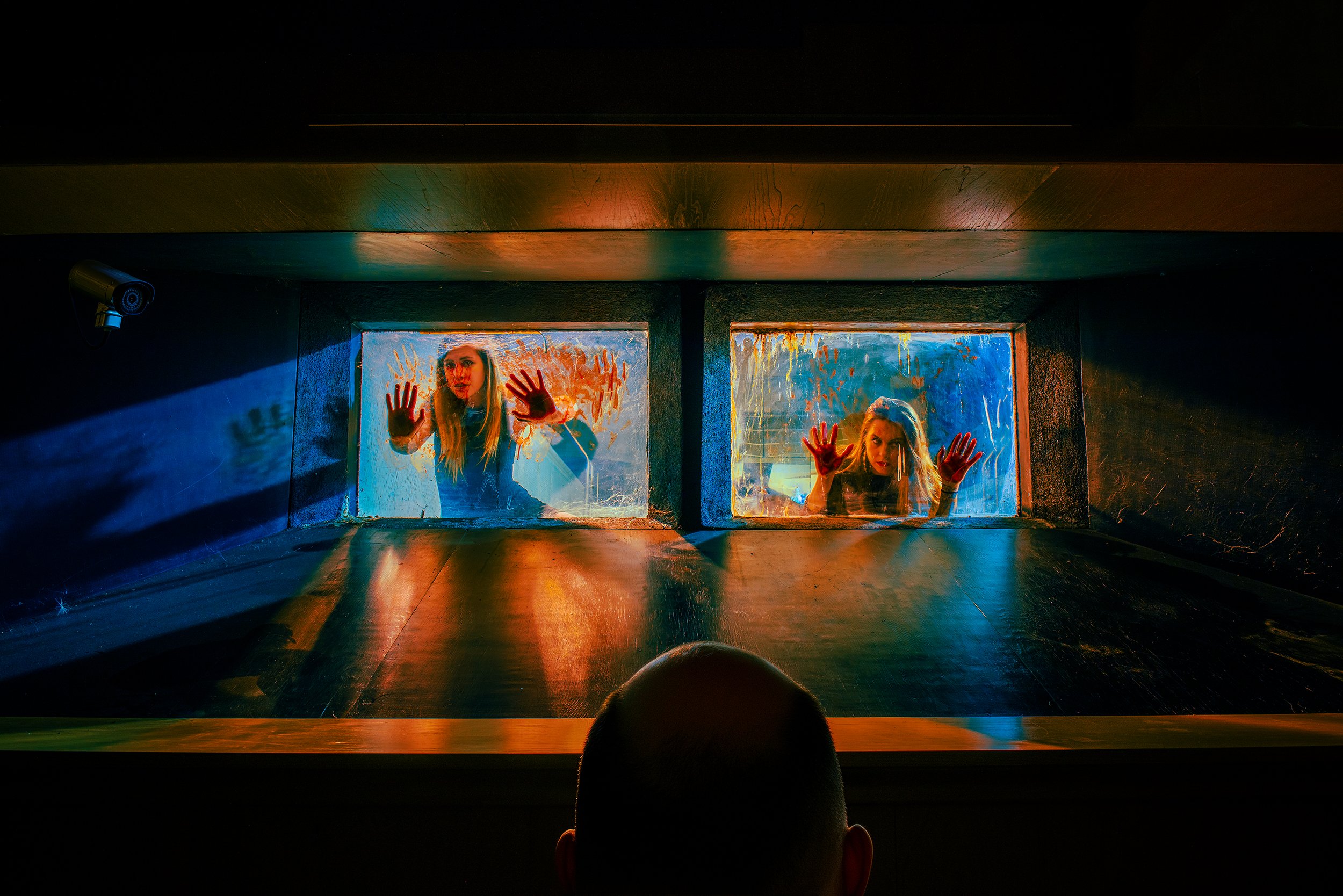 Two women looking through two adjacent glass windows, with their hands pressed against the glass, illuminated by colorful lights, in a dark room.