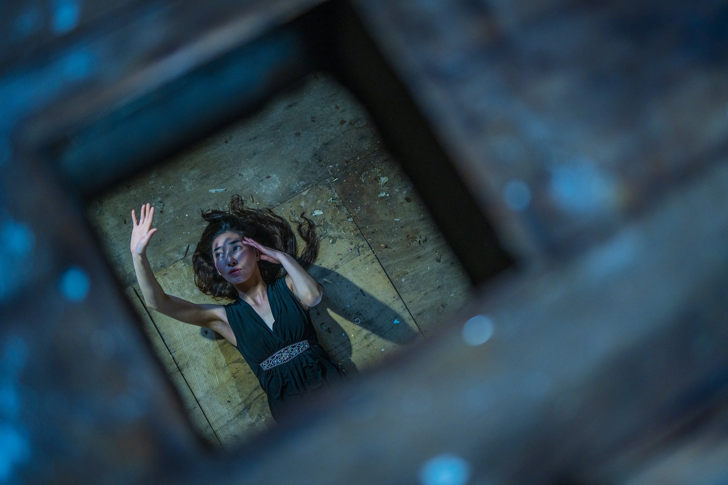 A woman lying on the ground with her hair spread around her head, seen through a frame or opening, with her hand raised near her face.