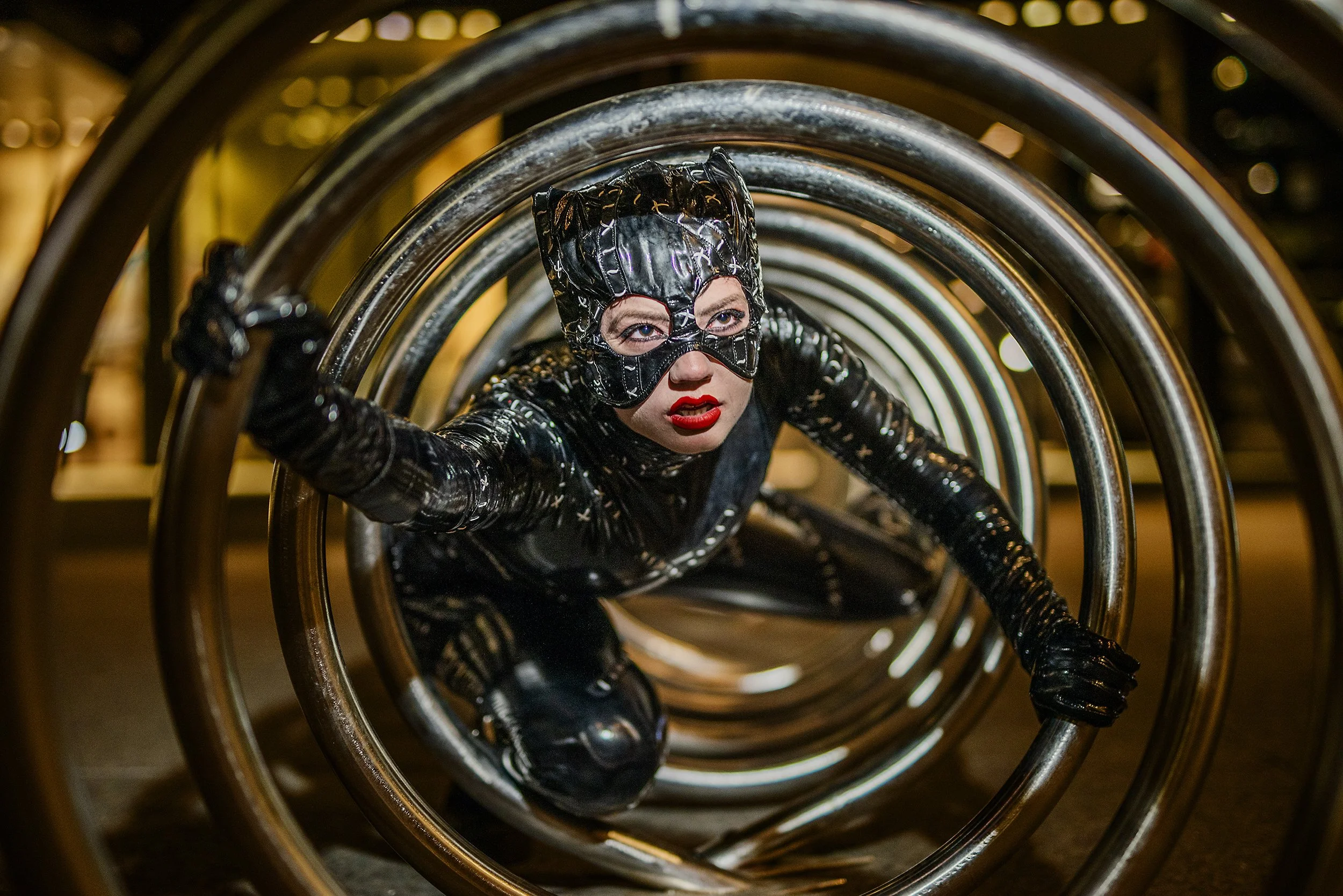 Person dressed as Catwoman crawling through metallic bike racks at night, wearing a shiny black costume, mask, and red lipstick.