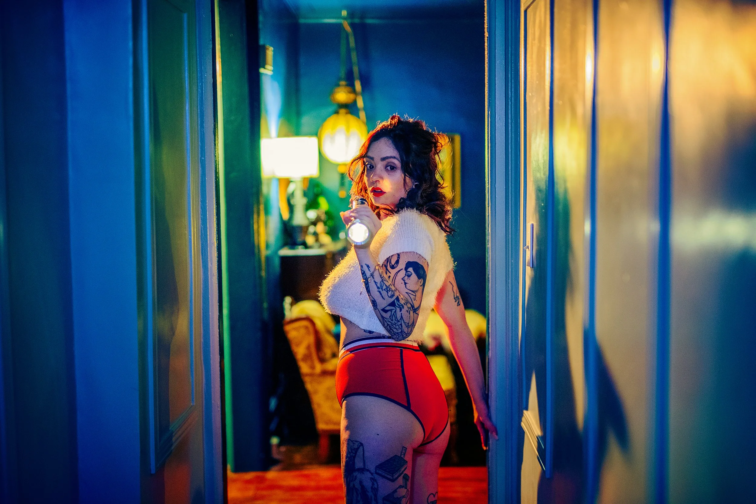 A woman with tattoos on her arms and legs, dark curly hair, wearing a white top and red pants, stands in a narrow hallway illuminated by colorful lights, holding a flashlight and looking over her shoulder.