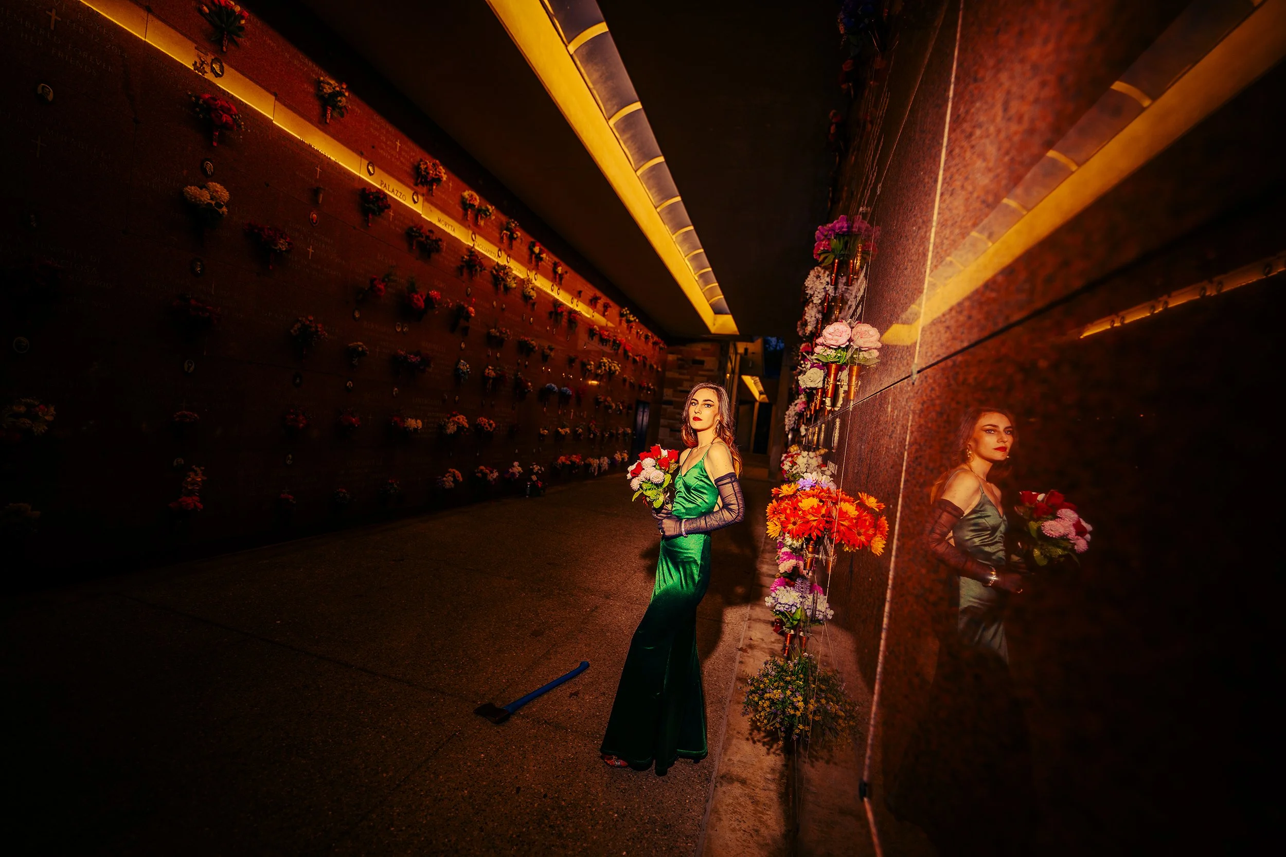 Two women in elegant dresses holding bouquets of flowers stand beside a reflective wall with floral arrangements. The scene is lit with warm lighting at night.