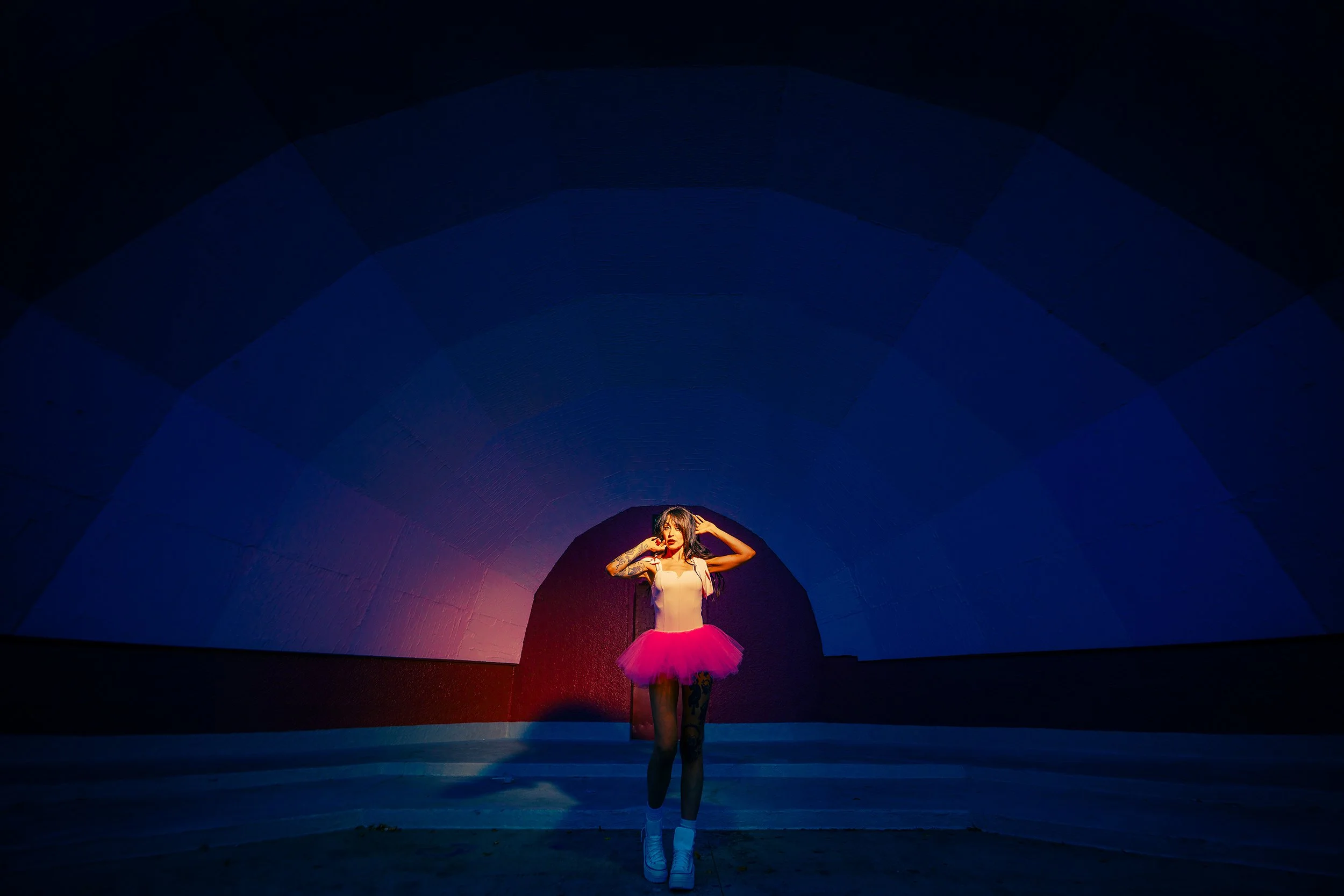 A woman in a pink tutu skirt and white top standing in a tunnel with colorful lighting, with the light illuminating her face and body.