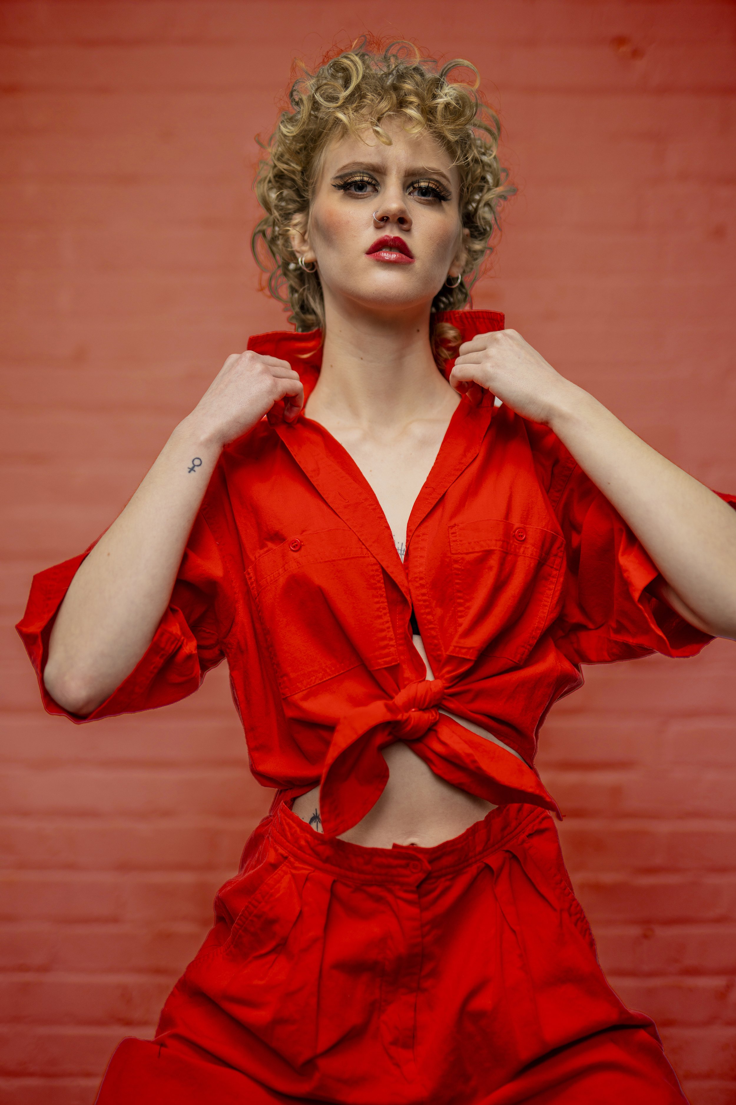 A woman with curly blonde hair, red lipstick, and eyeliner, wearing a red jacket tied at the waist, and matching red pants, poses against a red background.