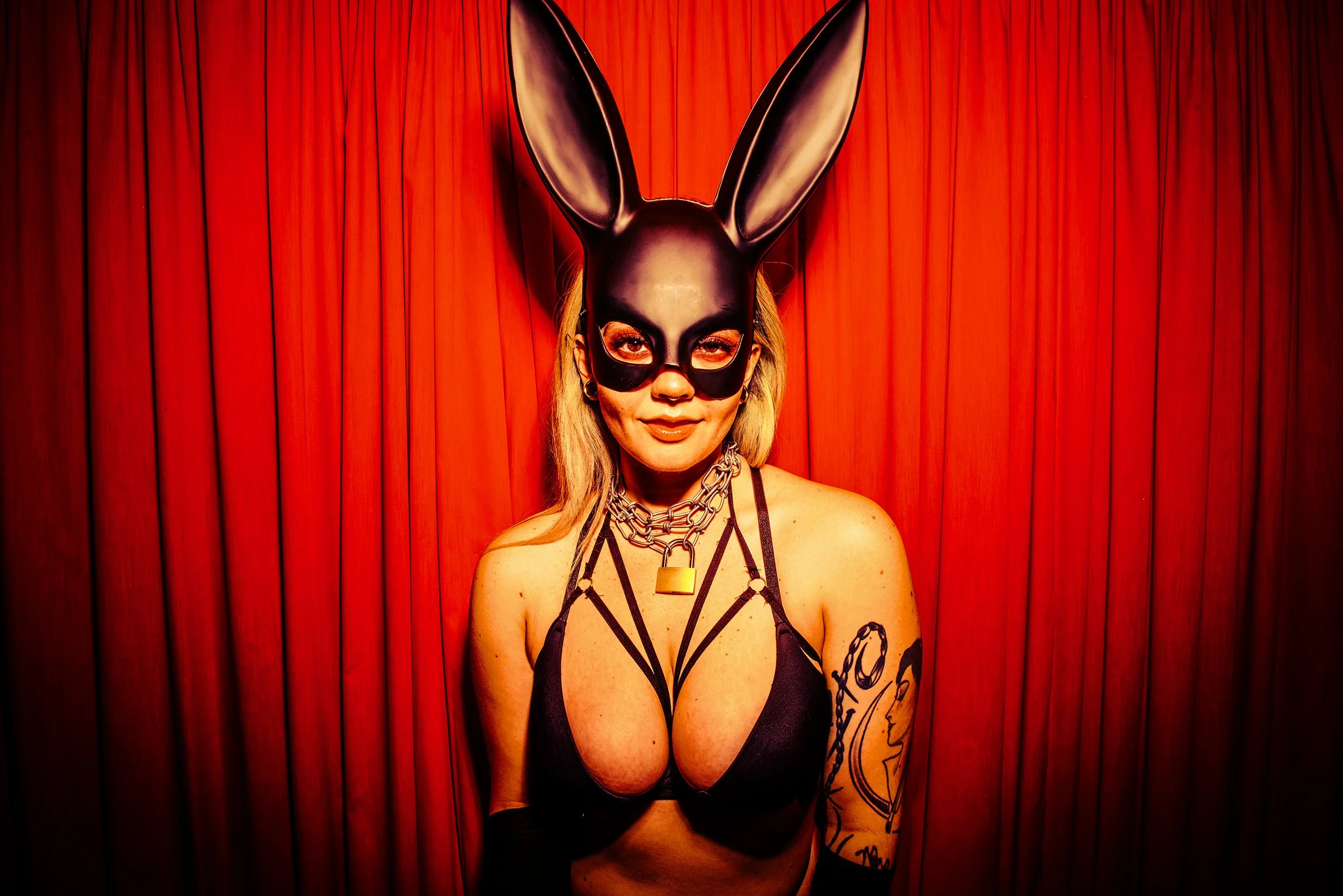 A woman wearing a black rabbit mask, glasses, and jewelry, standing against a red curtain.