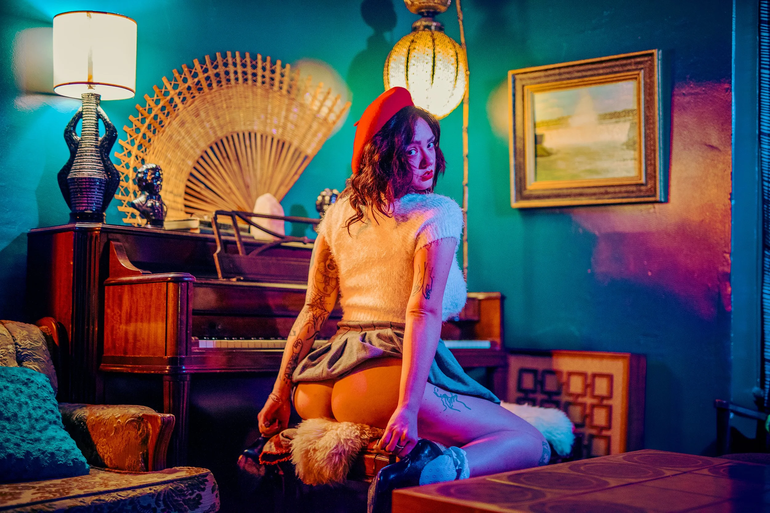 A woman with dark curly hair and tattoos on her arm, wearing a red beret and beige top, kneeling on a fluffy rug in a colorful living room with teal walls, a wooden piano, a framed landscape painting, and various lamps and decorative items.