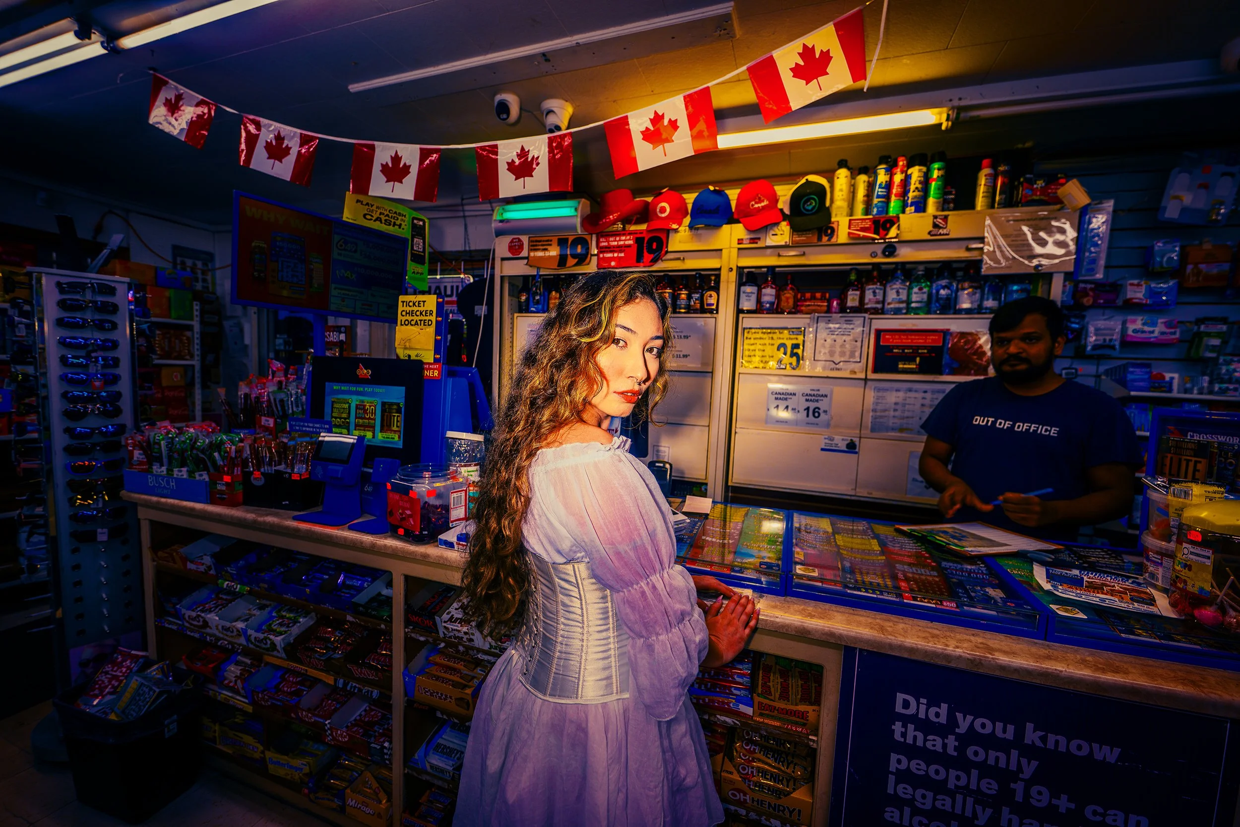 A woman with long wavy hair and light-colored clothing standing at a checkout counter in a convenience store, with a man in a dark shirt behind the counter. The store is decorated with Canadian flags and various products on shelves.