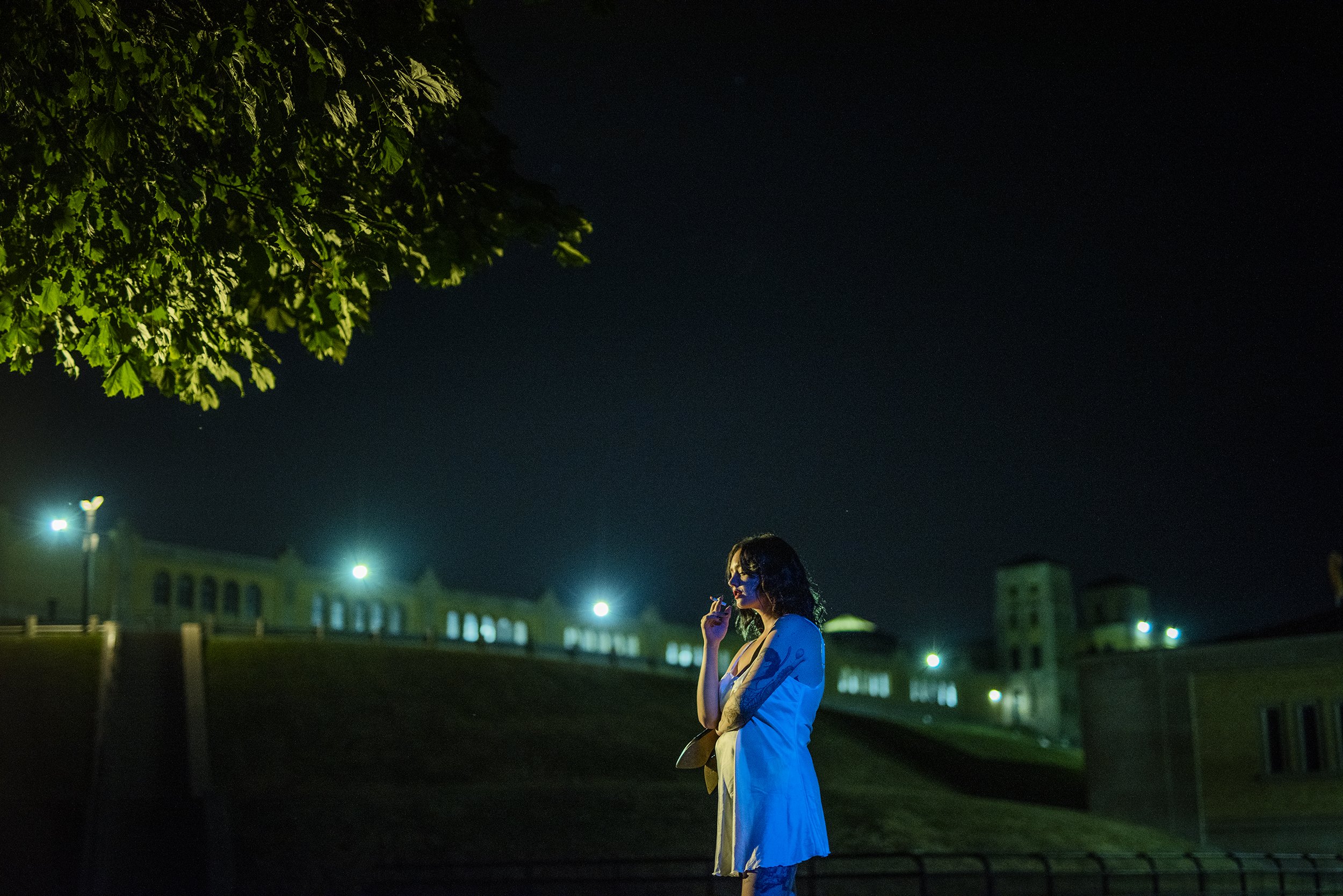 A woman in a light-colored dress standing outside at night, smoking a cigarette, with illuminated buildings and bright streetlights in the background.