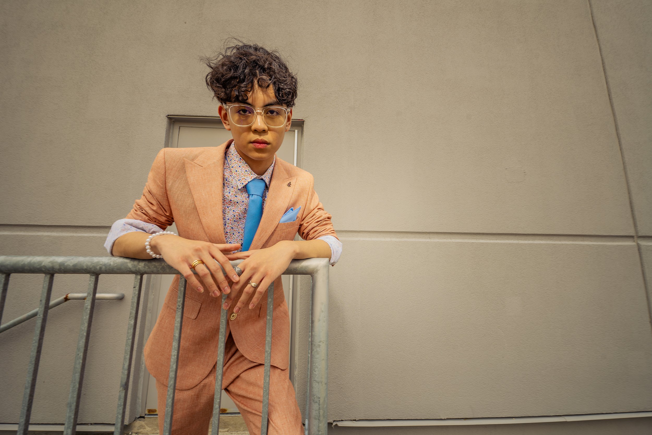 A young man with curly hair and glasses leaning on a metal railing, wearing an orange suit, a colorful shirt, a blue tie, and accessories, standing in front of a plain beige wall.