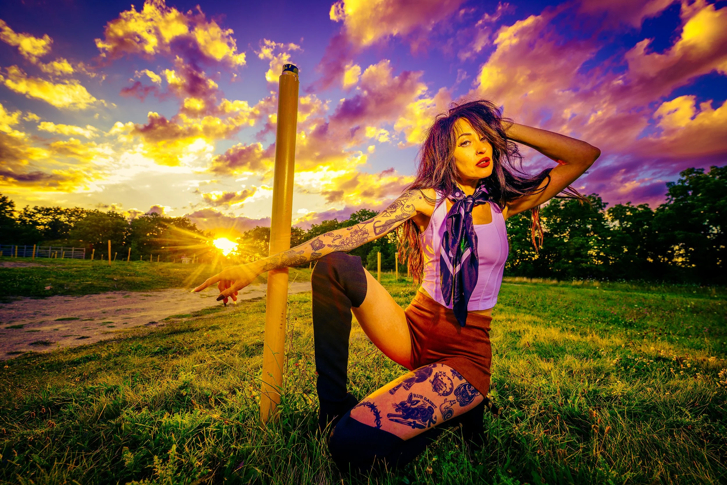 A woman with tattoos in a purple top and red pants poses outdoors during sunset, reaching out to touch a pole with a vibrant sky filled with purple and yellow clouds in the background.