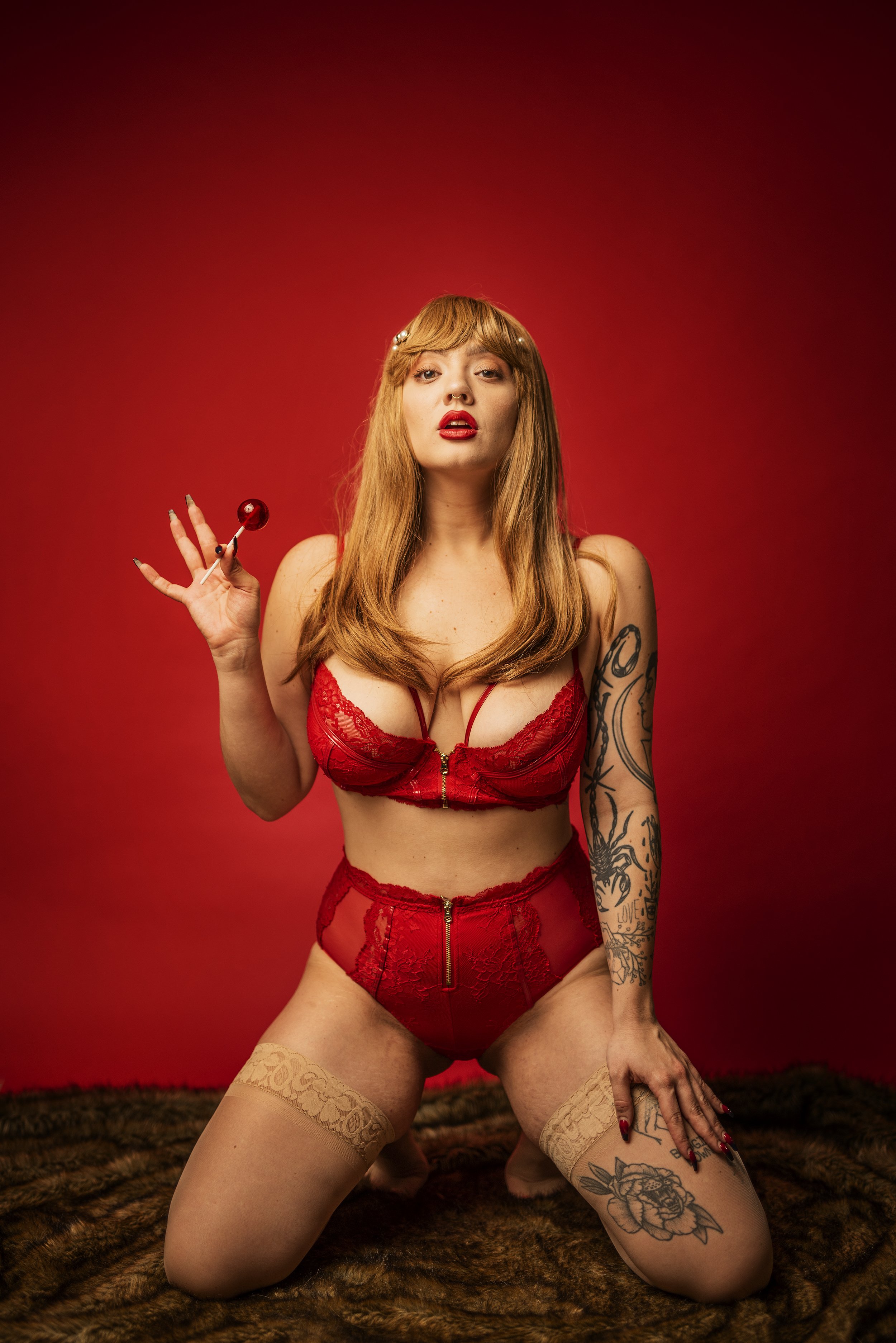 A woman with long red hair, red lipstick, and tattoos wearing red lingerie, kneeling on a furry surface against a red background, holding a red lollipop.