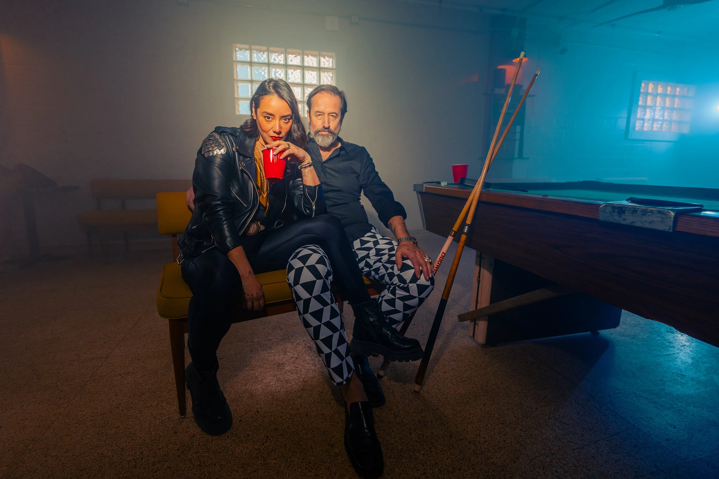 Two people sitting on a yellow bench in a foggy room, one woman drinking from a red cup, the other man with a beard, wearing patterned pants and holding a pool cue, near a pool table.