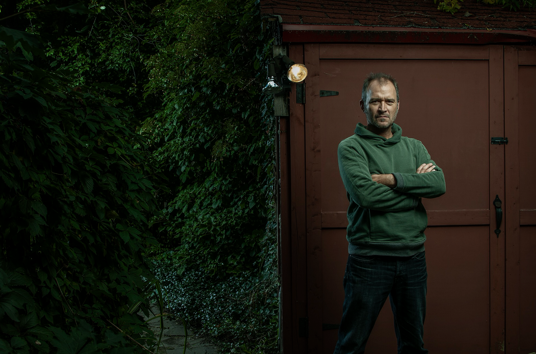 A man with crossed arms standing in front of a red shed, with green foliage on the left side of the image.