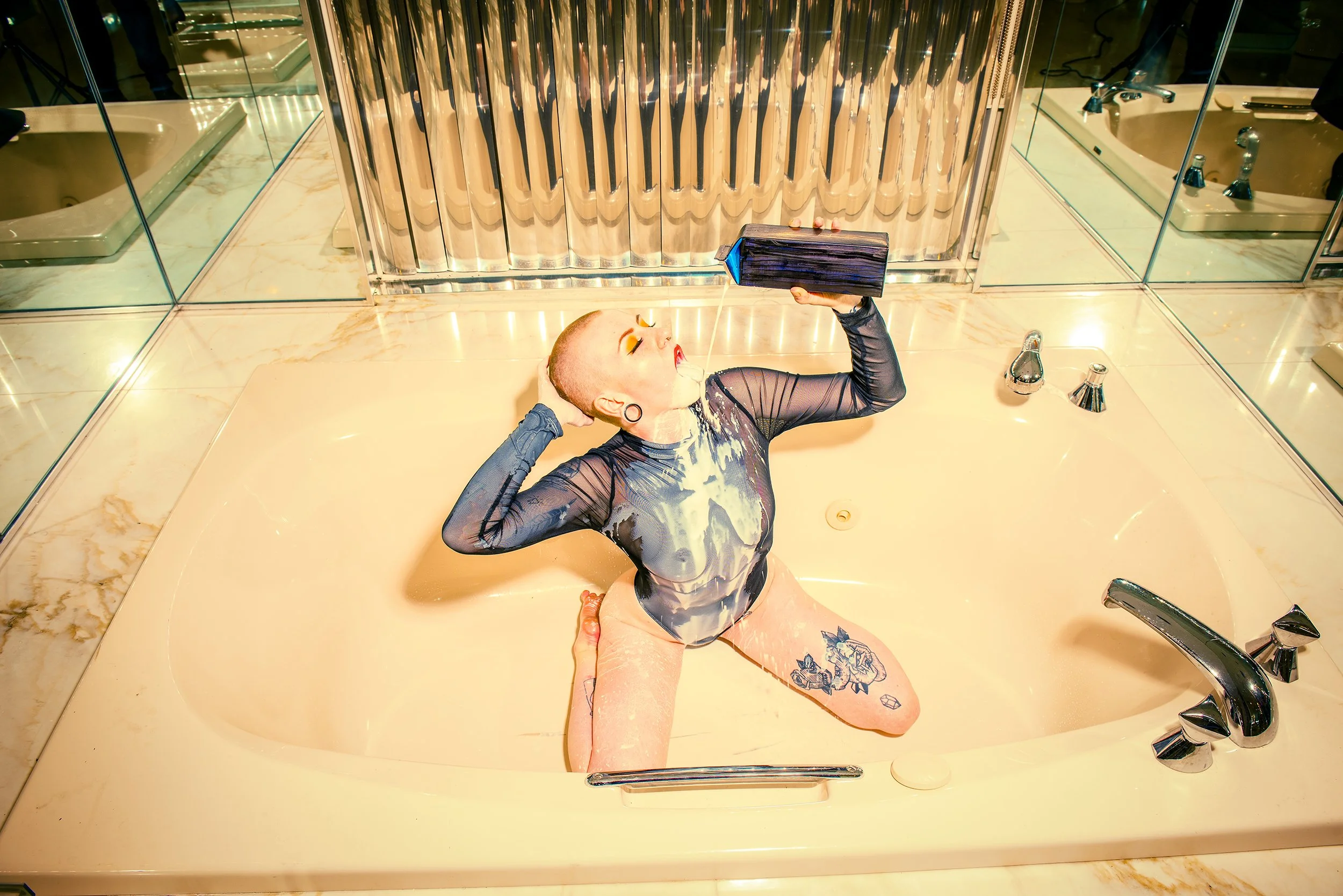 A person with a shaved head and tattoos on their leg sitting in a bathtub while someone else pours water over their face.