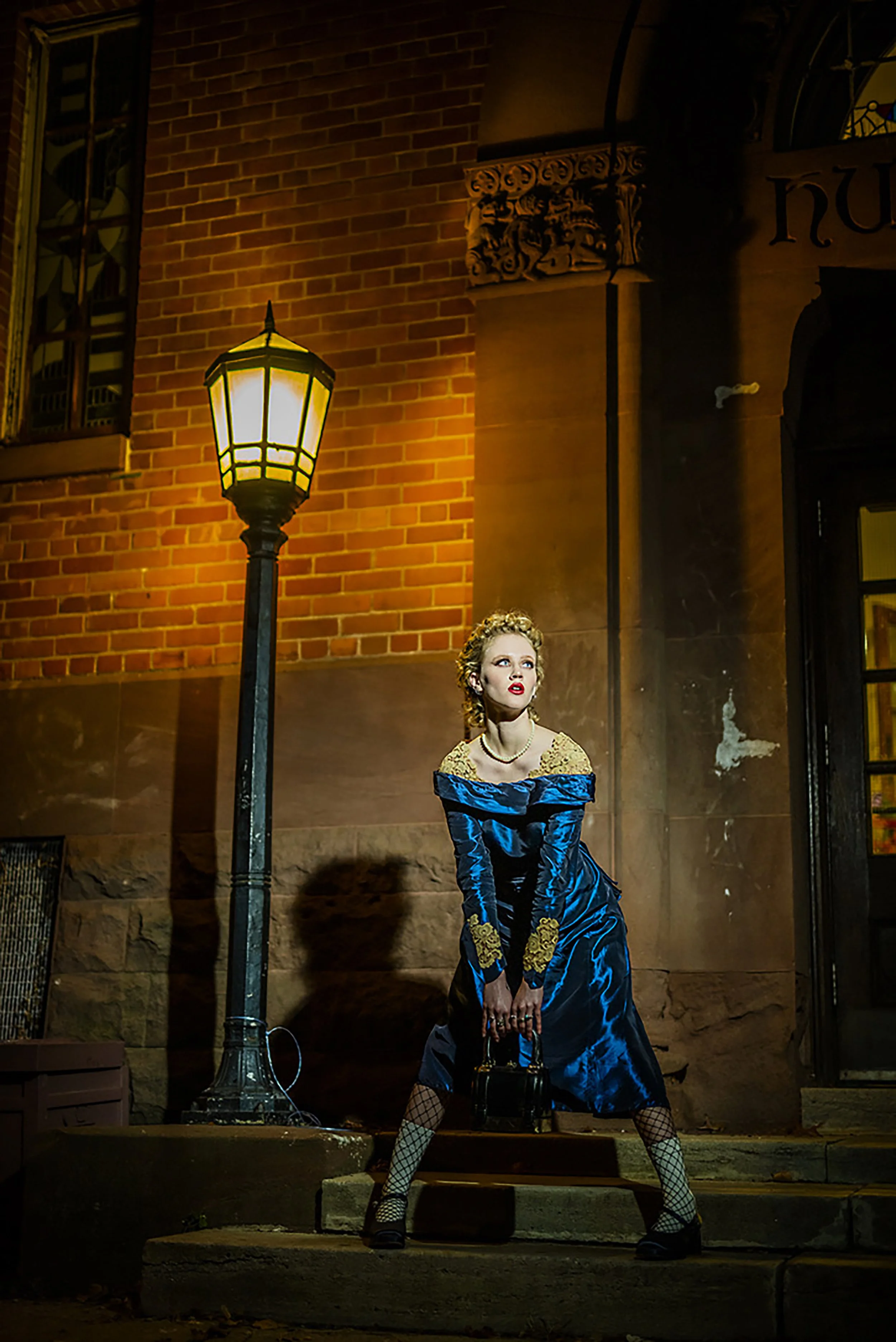 A woman dressed in vintage style with a blue satin gown, yellow lace accents, fishnet stockings, and black shoes, posing on a city street at night near a vintage streetlamp, with brick buildings in the background.