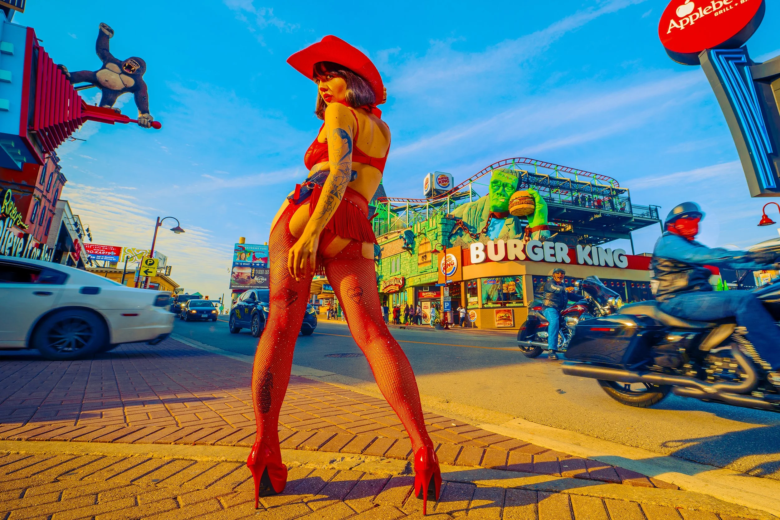 A woman dressed in red fishnet tights, red skirt, red top, and red high heels with a red hat, standing on a city street during sunset. The background features a Burger King restaurant, a large Frankenstein figure, and a sign with an ape holding a ham
