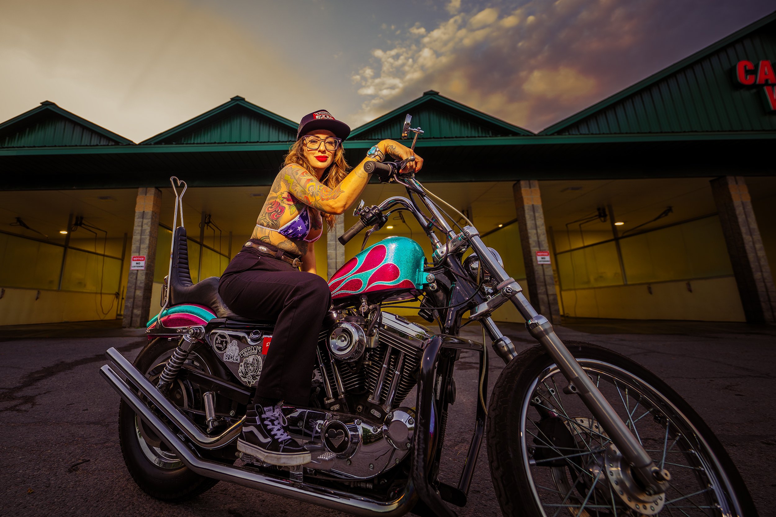 A woman with tattoos and glasses sitting on a colorful motorcycle in front of a building at sunset.
