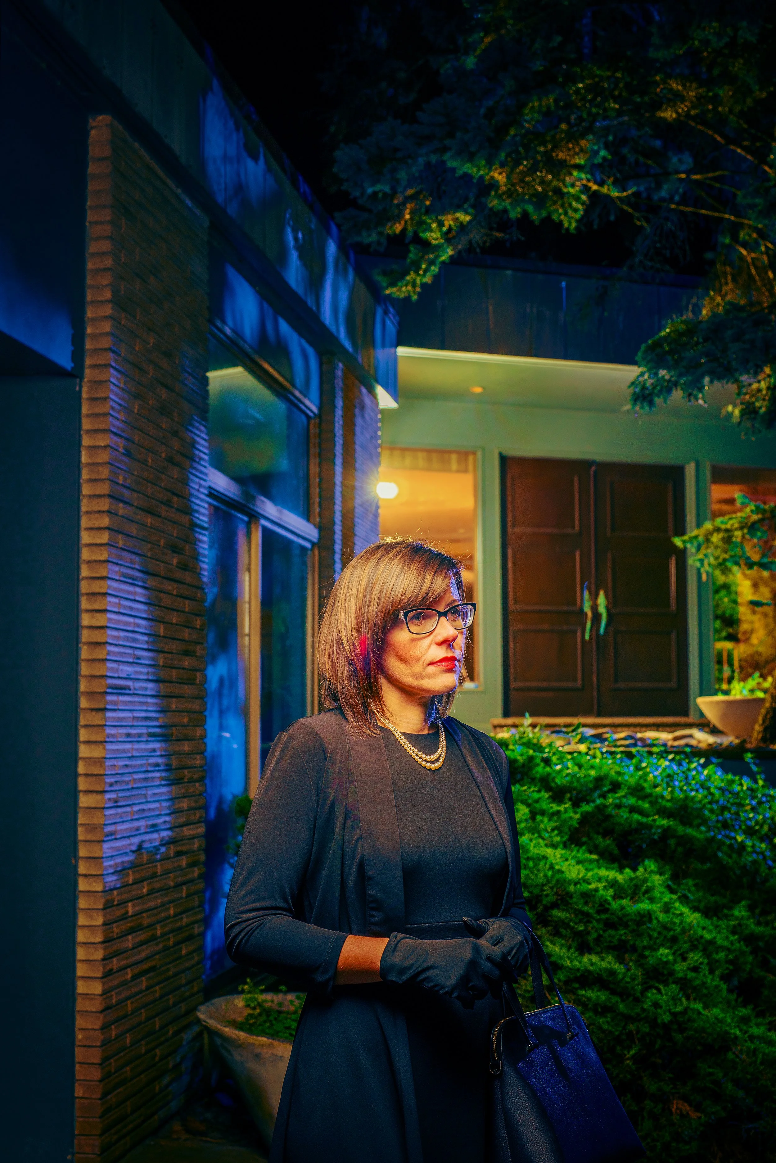 A woman with shoulder-length brown hair, wearing glasses, a black dress, a pearl necklace, and black gloves, standing outside at night near a house with a brick wall, large windows, and a green door, illuminated by outdoor lighting, with trees and gr