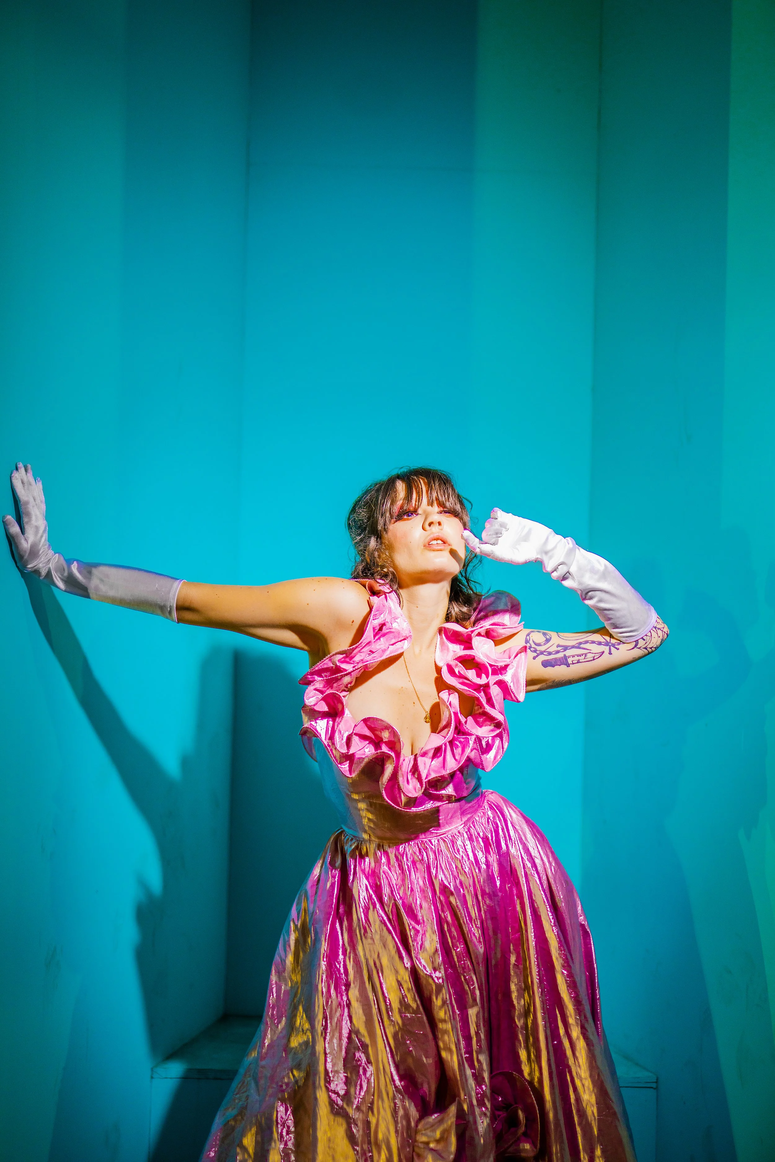 A woman wearing a pink and gold ruffled dress and white gloves is posing with one hand on her face and the other pressed against a teal wall.