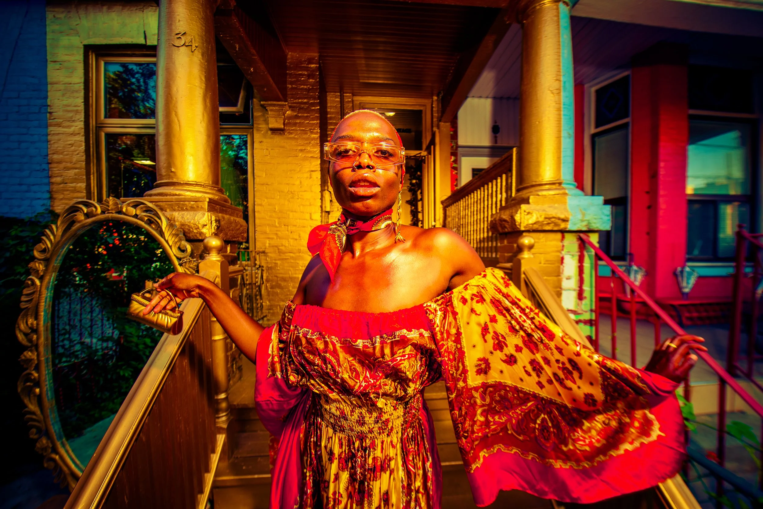 Person wearing glasses and a colorful off-shoulder dress standing in front of a porch with brick pillars and a railing, with vibrant multicolored lighting effects.