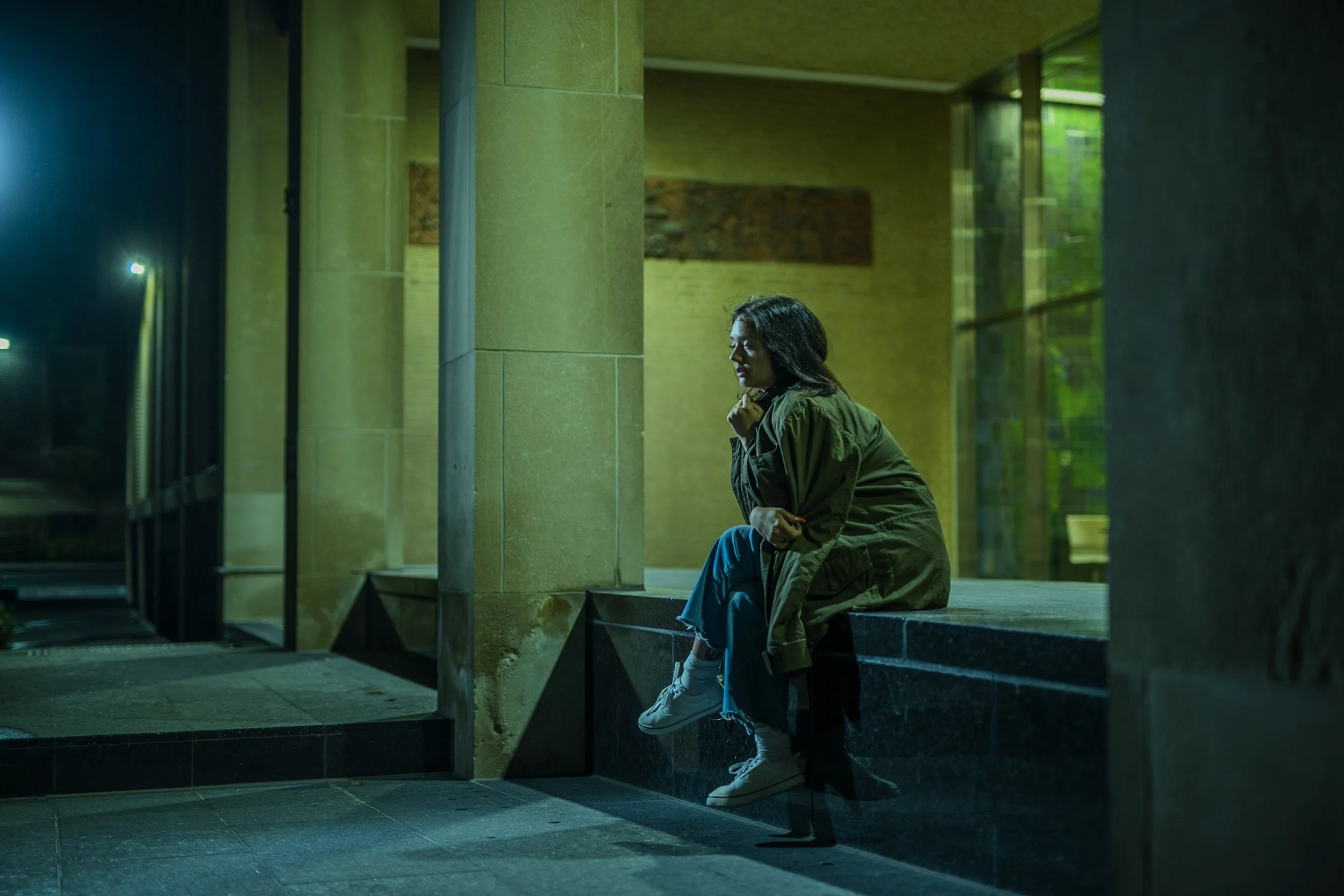 A woman sitting alone outside a building at night, resting her chin on her hand, illuminated by greenish light through large windows.