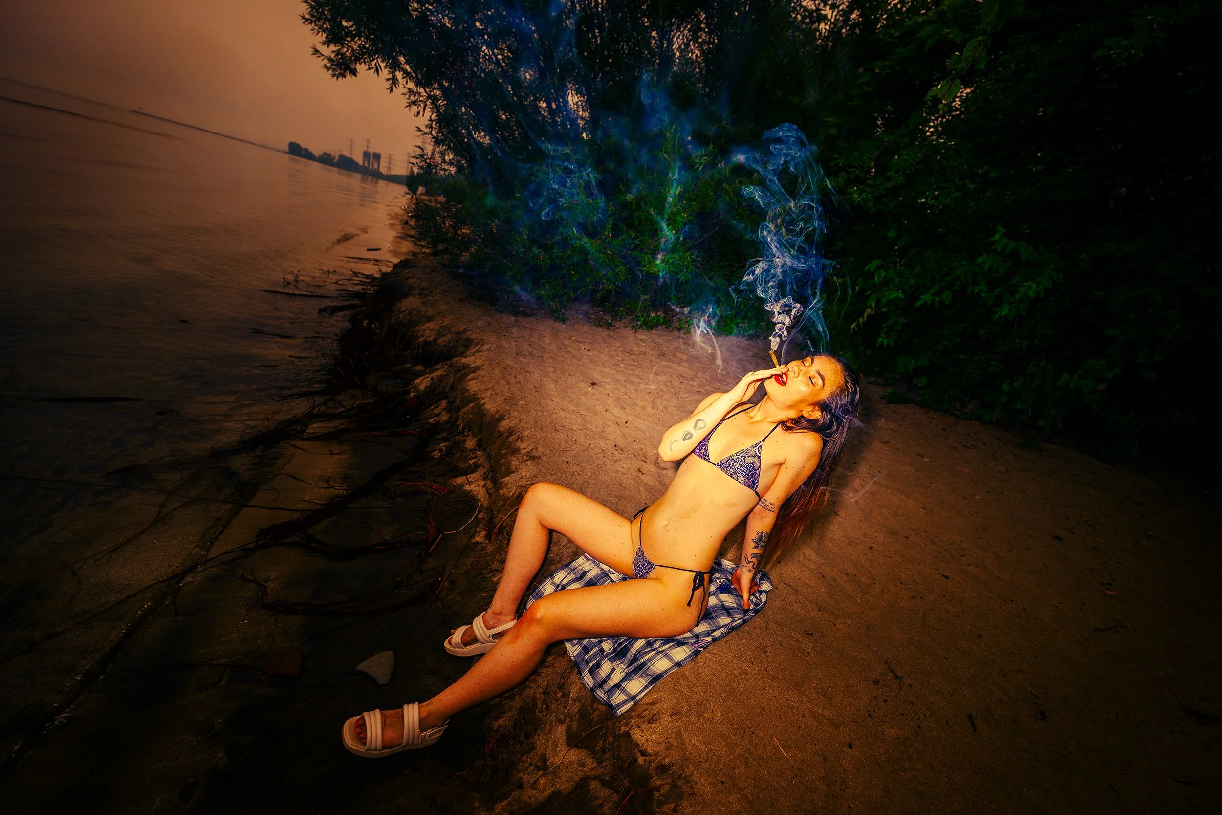 A woman lying on the ground near the water's edge, smoking, with smoke rising above her head. She is wearing a swimsuit, has tattoos, and is surrounded by dark trees and a cloudy sky.