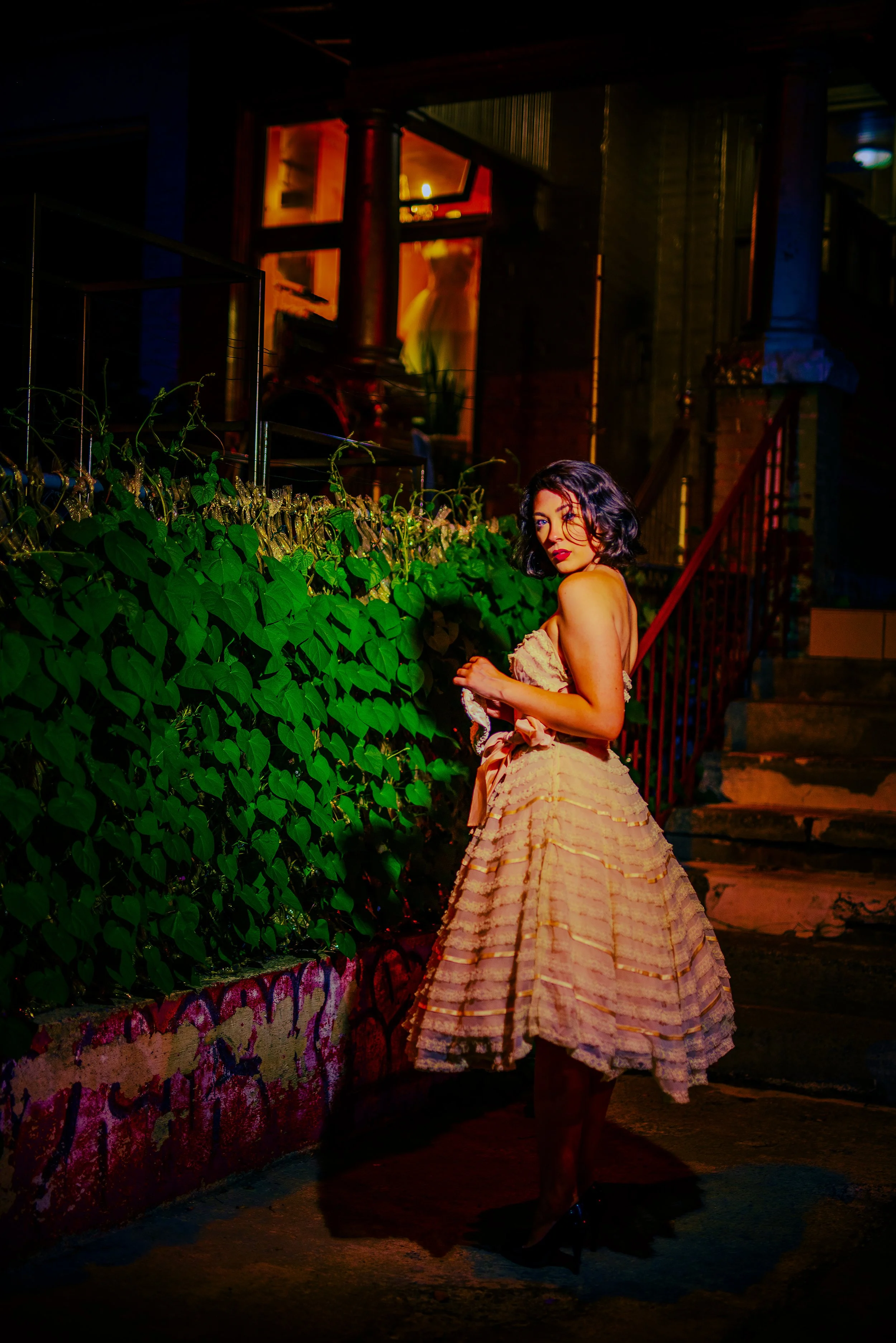 A woman with black curly hair and red lipstick wearing a yellow ruffled dress with a full skirt stands outdoors at night in front of a green leafy bush and a brick wall, illuminated by colorful lighting.