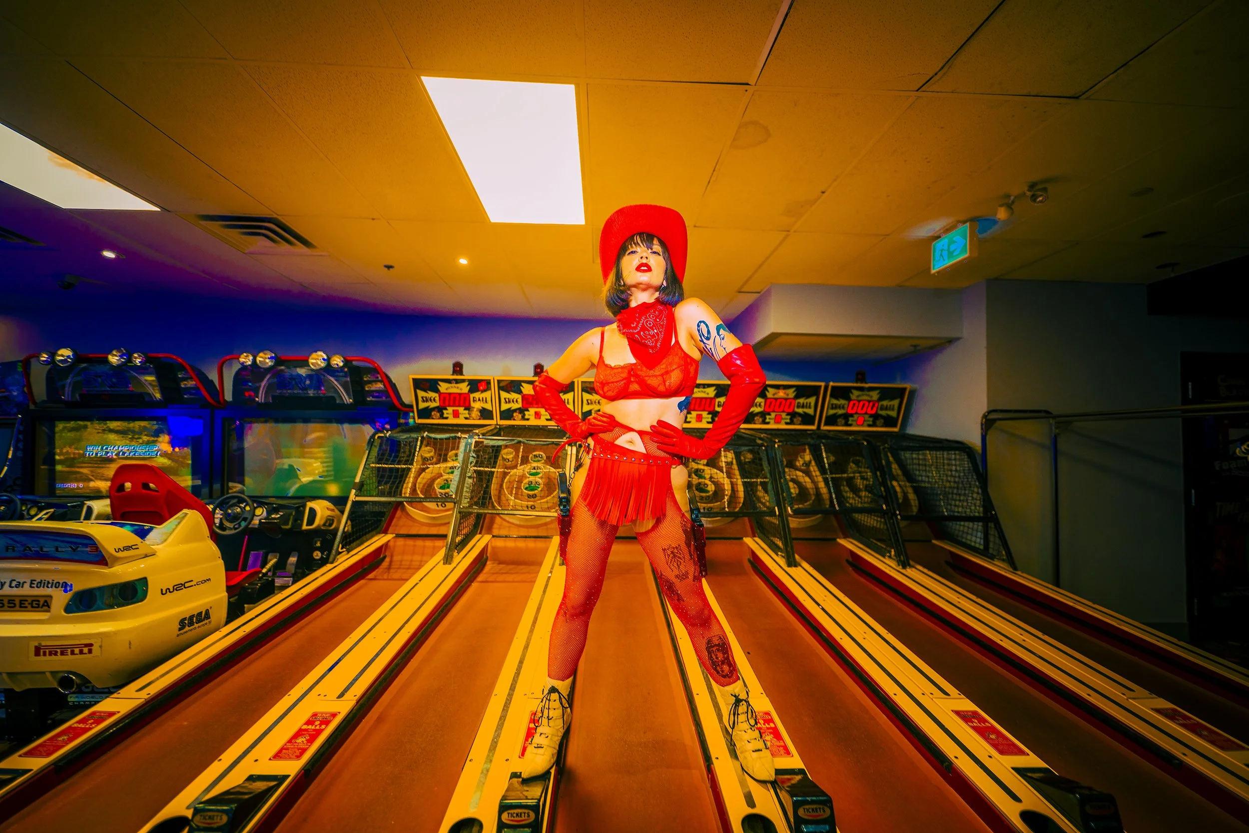 A woman dressed in red lingerie, fishnets, and cowboy boots standing on a racing arcade game lane at an indoor arcade, with slot machine-like games in the background.