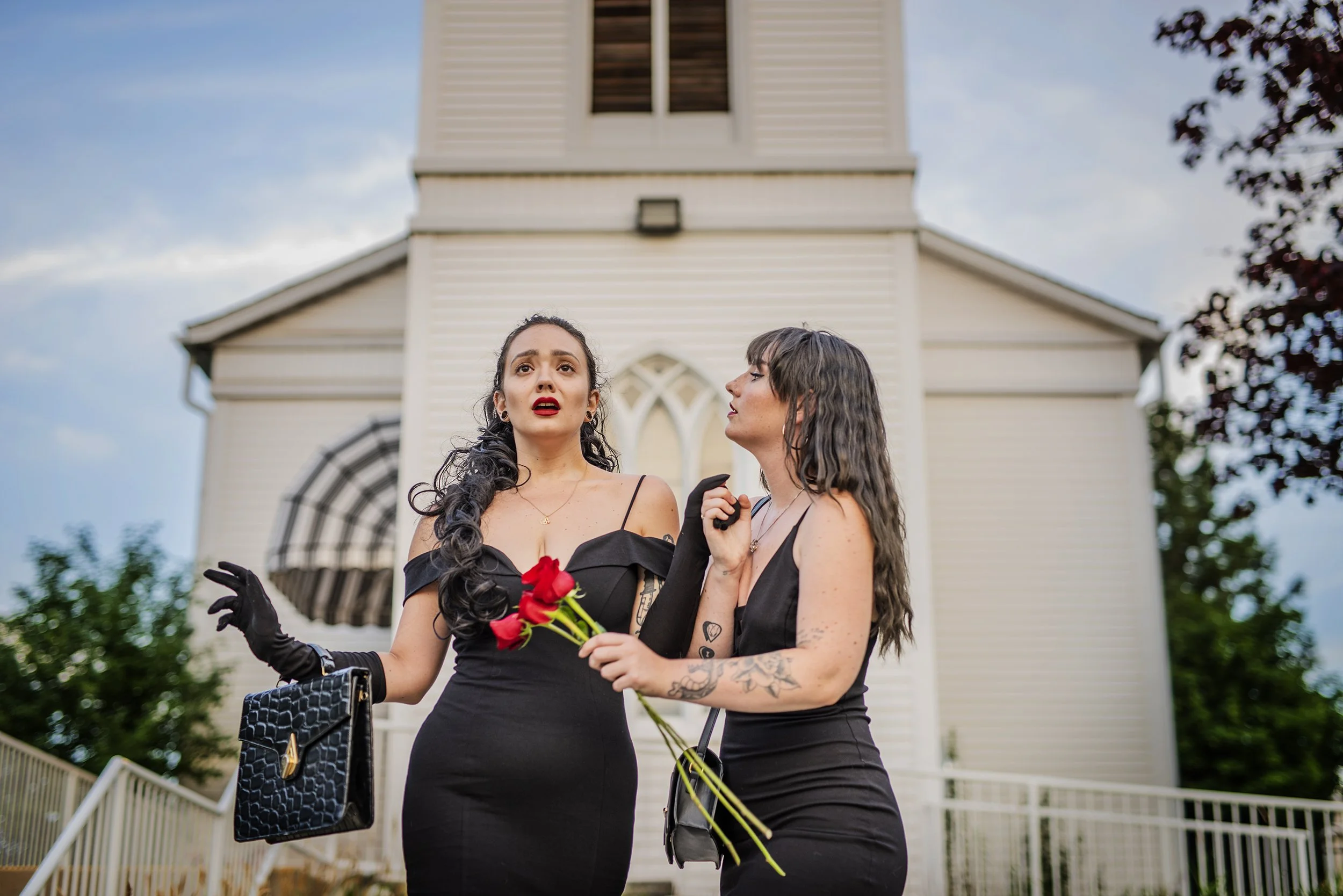 Two women in black dresses standing in front of a church, one holding a bouquet of red flowers and the other a black purse, with emotional expressions on their faces.