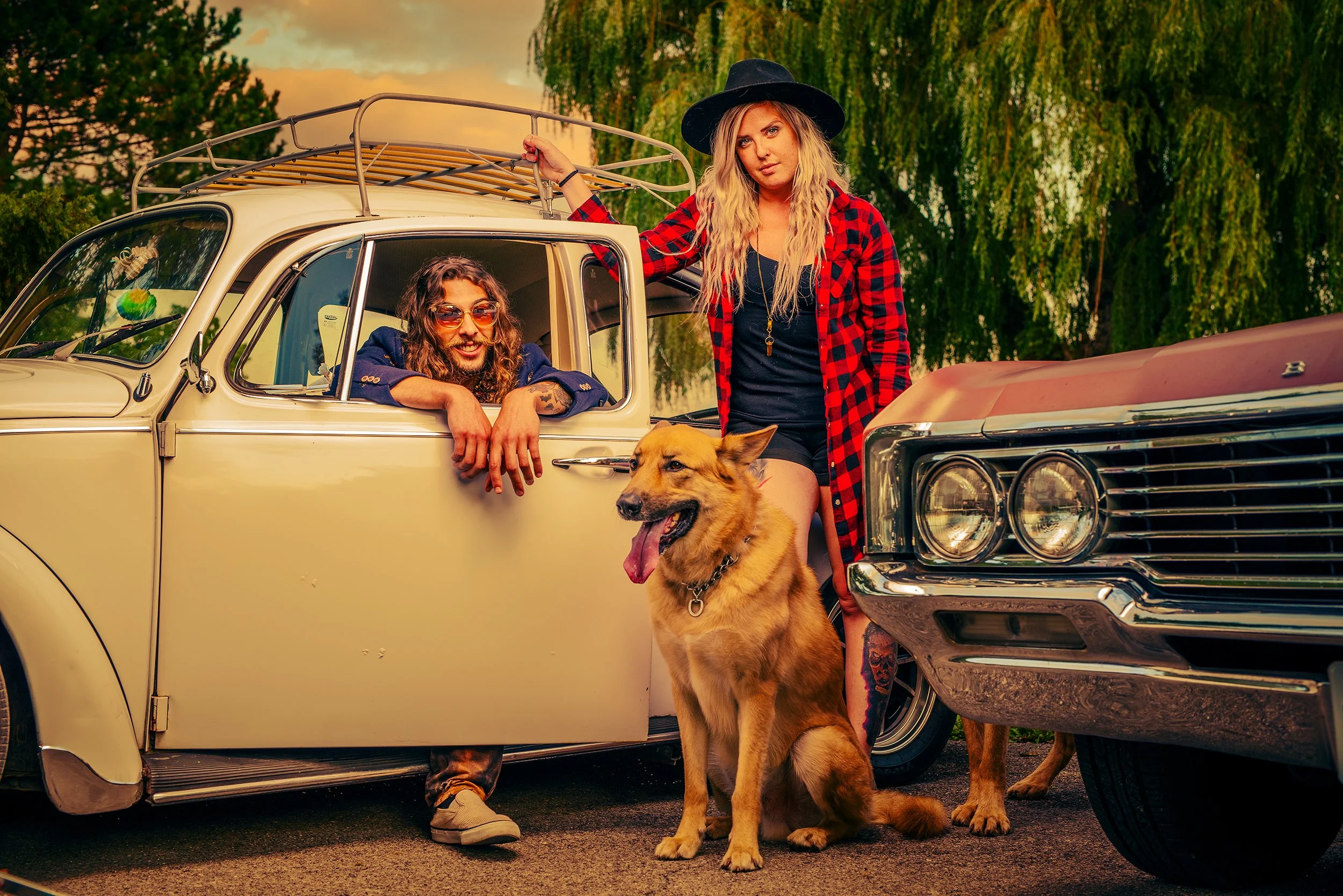 Two young women with long hair and casual clothing posing with a vintage cream-colored car outdoors during sunset. One is leaning on the car's roof holding onto a rack, the other is kneeling in front of the car, with a German Shepherd dog sitting bes