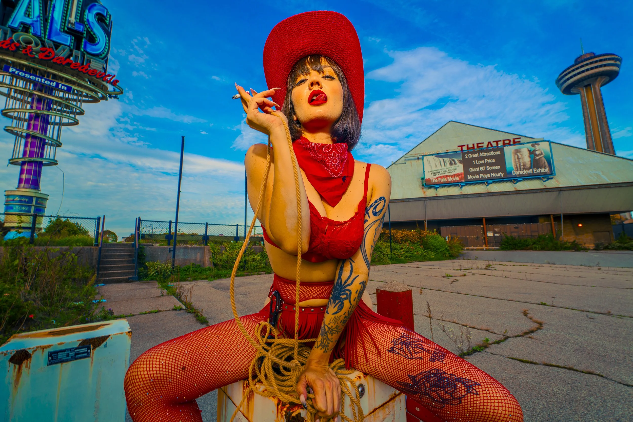 A woman dressed in red fishnet clothing and a red hat, sitting on a box outdoors, holding a cigarette and posing with a confident expression in front of an abandoned amusement park with a theater sign and giant clown and tower in the background.