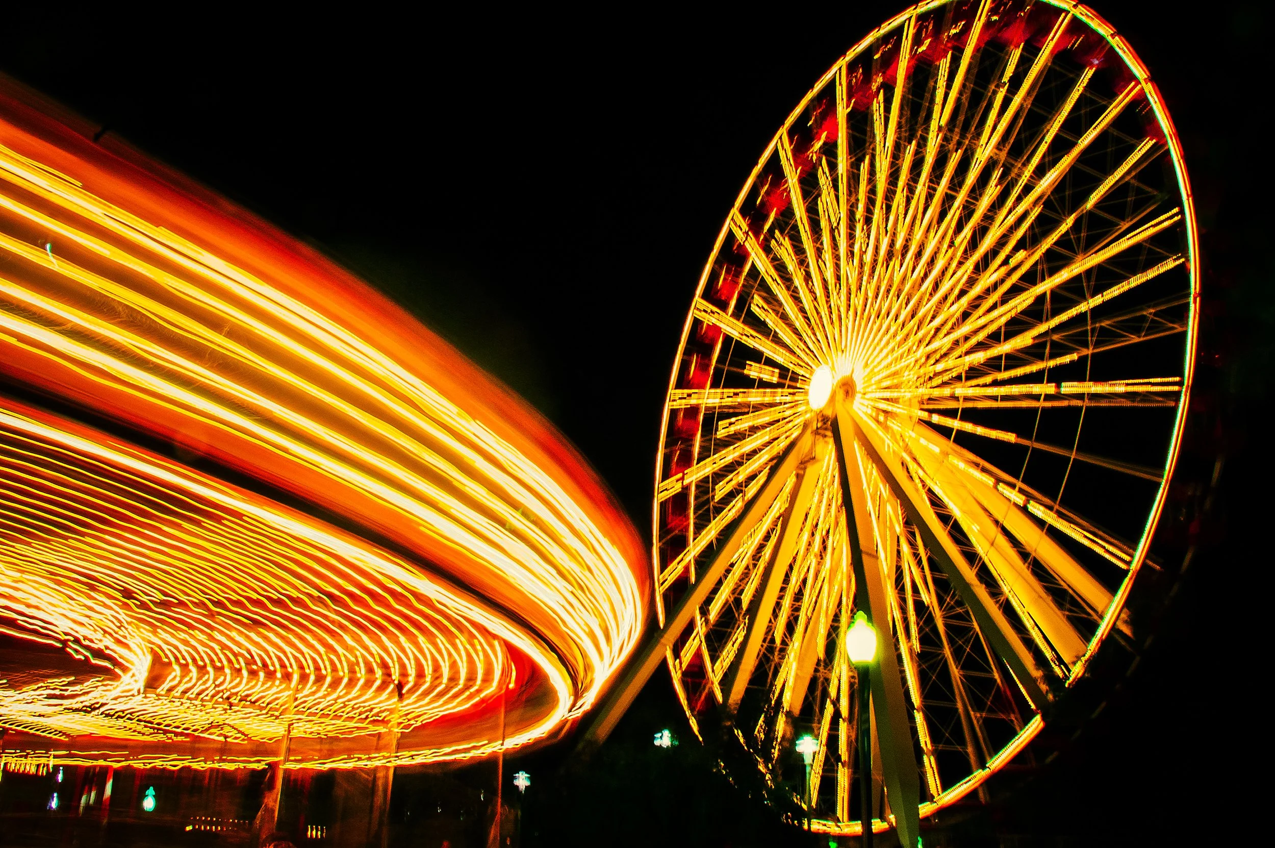 A brightly lit Ferris wheel at night, with streaks of colorful lights from the moving amusement park rides.
