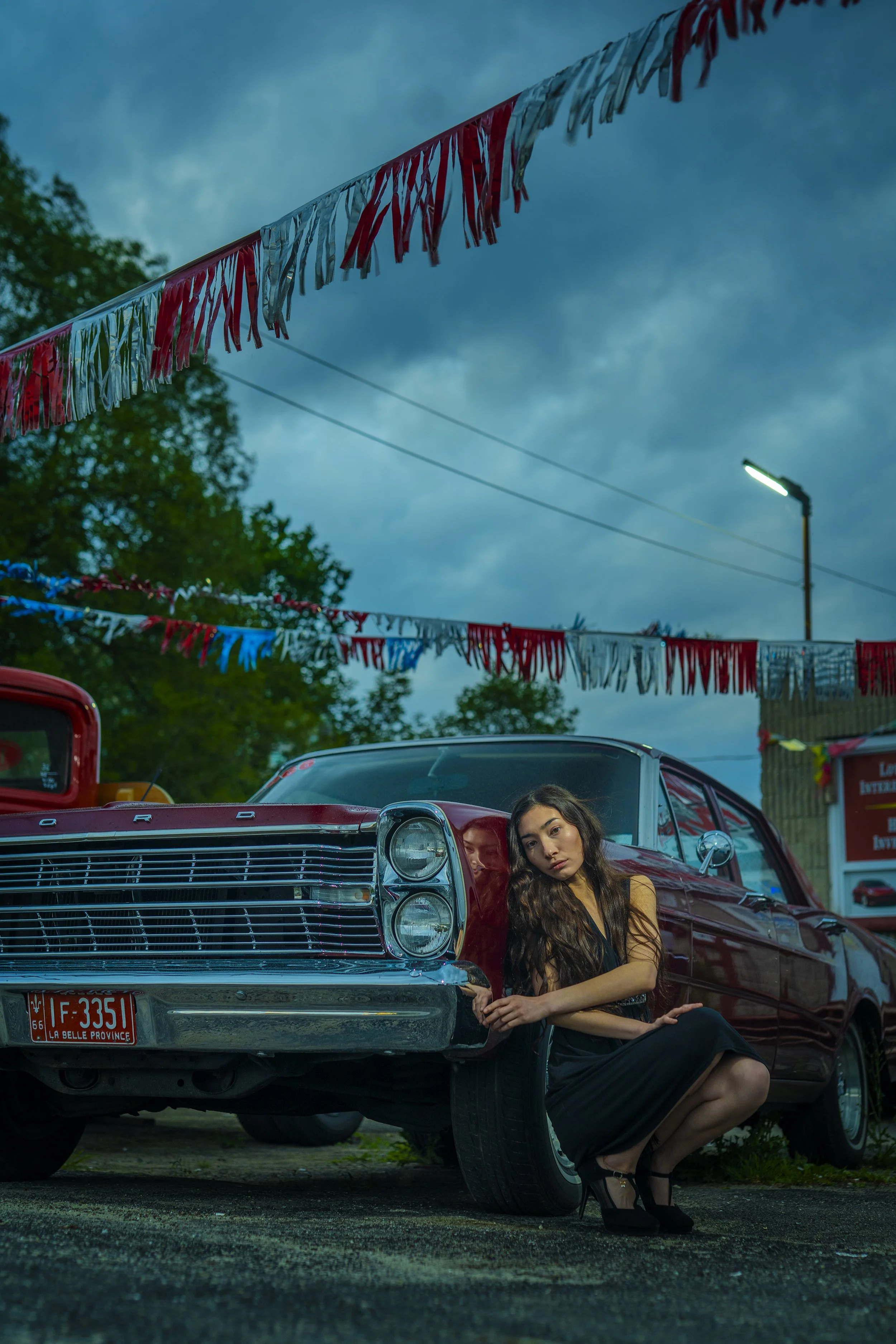 A young woman with long, dark hair, dressed in black, sitting beside a vintage red car with a low profile, to the right of the image. She is crouching with her arms resting on her knees and her head tilted slightly, looking at the camera with a serio