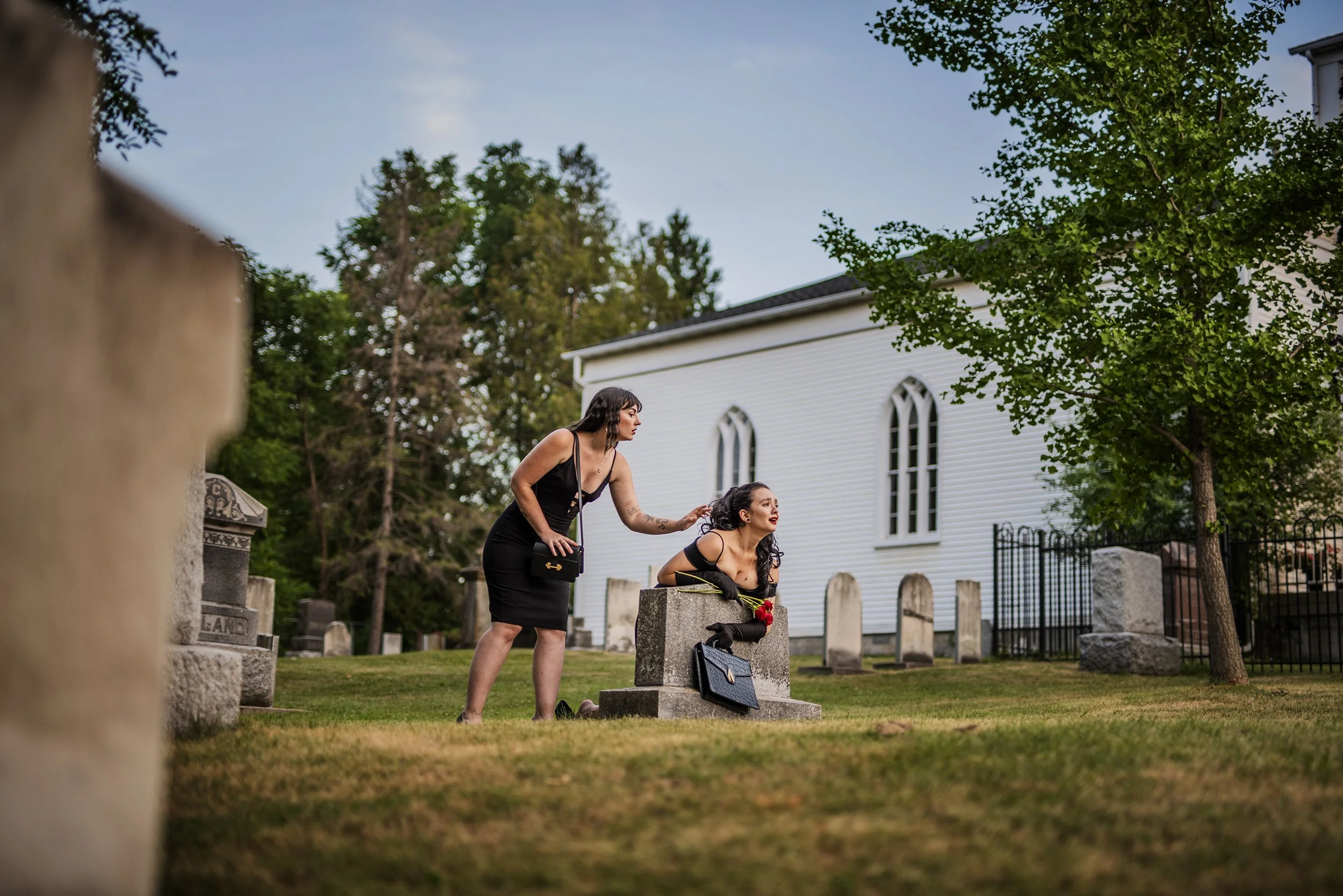 Two women in black dresses in a cemetery, one woman is kneeling on a tombstone holding a red flower, while the other woman is standing and touching her hair. A white church with arched windows and a black fence are in the background, with trees and a