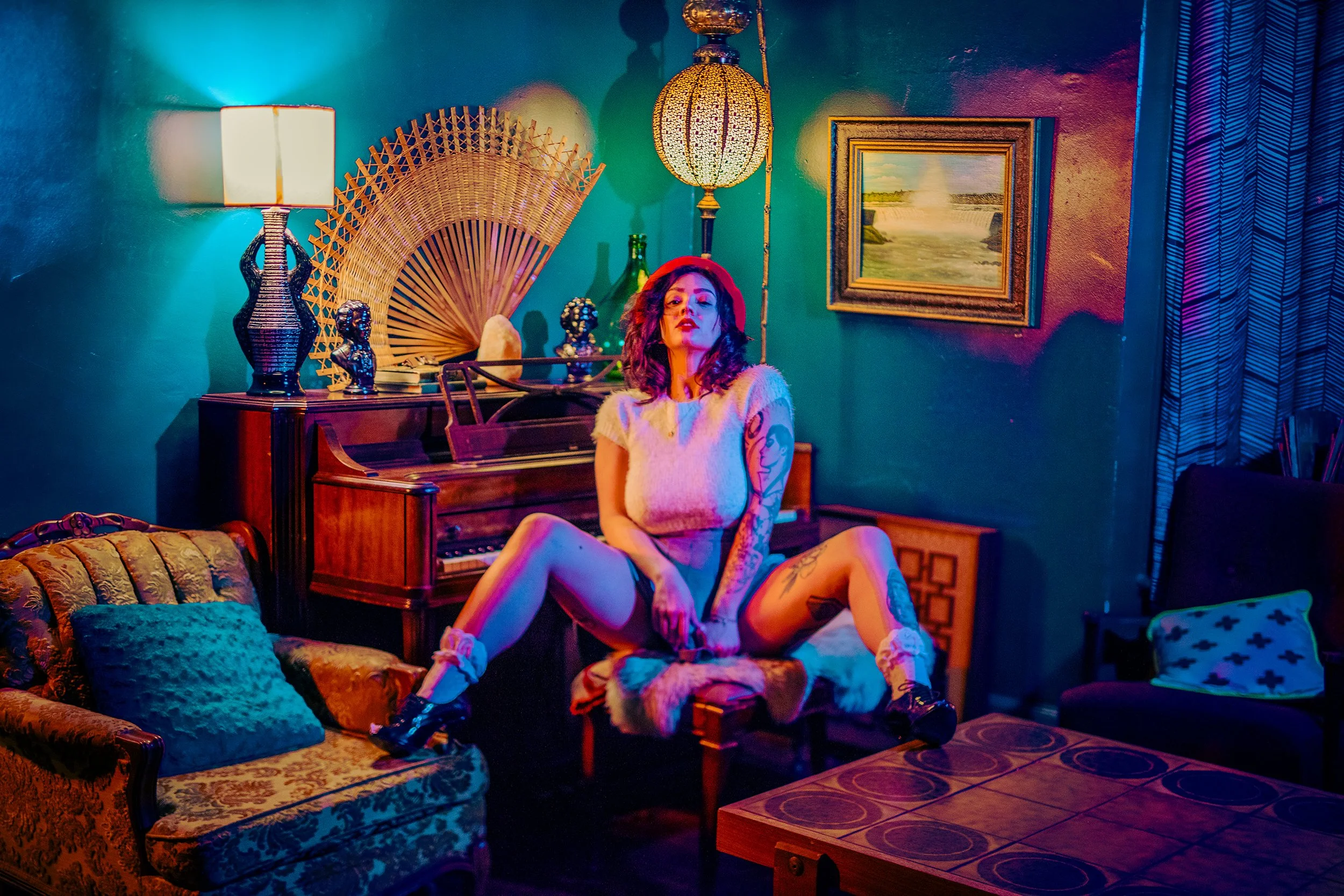 A woman with tattoos and curly hair, wearing a white top and shorts, sitting on a patterned sofa in a colorful, dimly lit room with eclectic decor including lamps, sculptures, paintings, and vintage furniture.