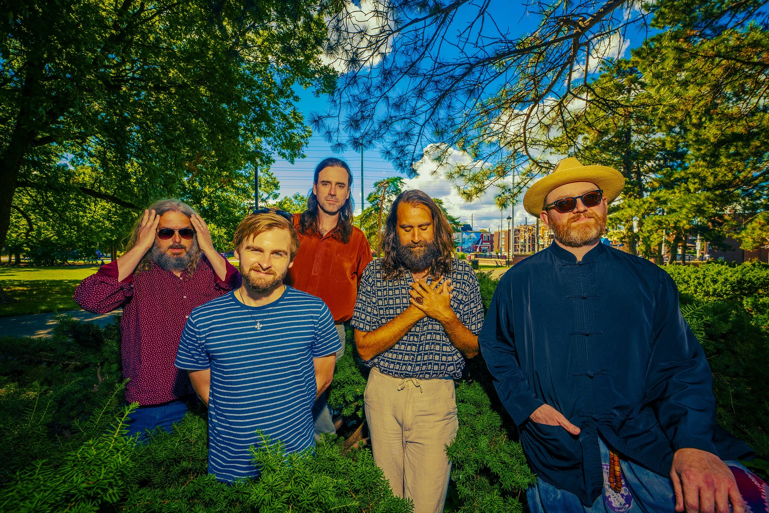 Five men standing together outdoors under trees on a sunny day, posing for a group photo.