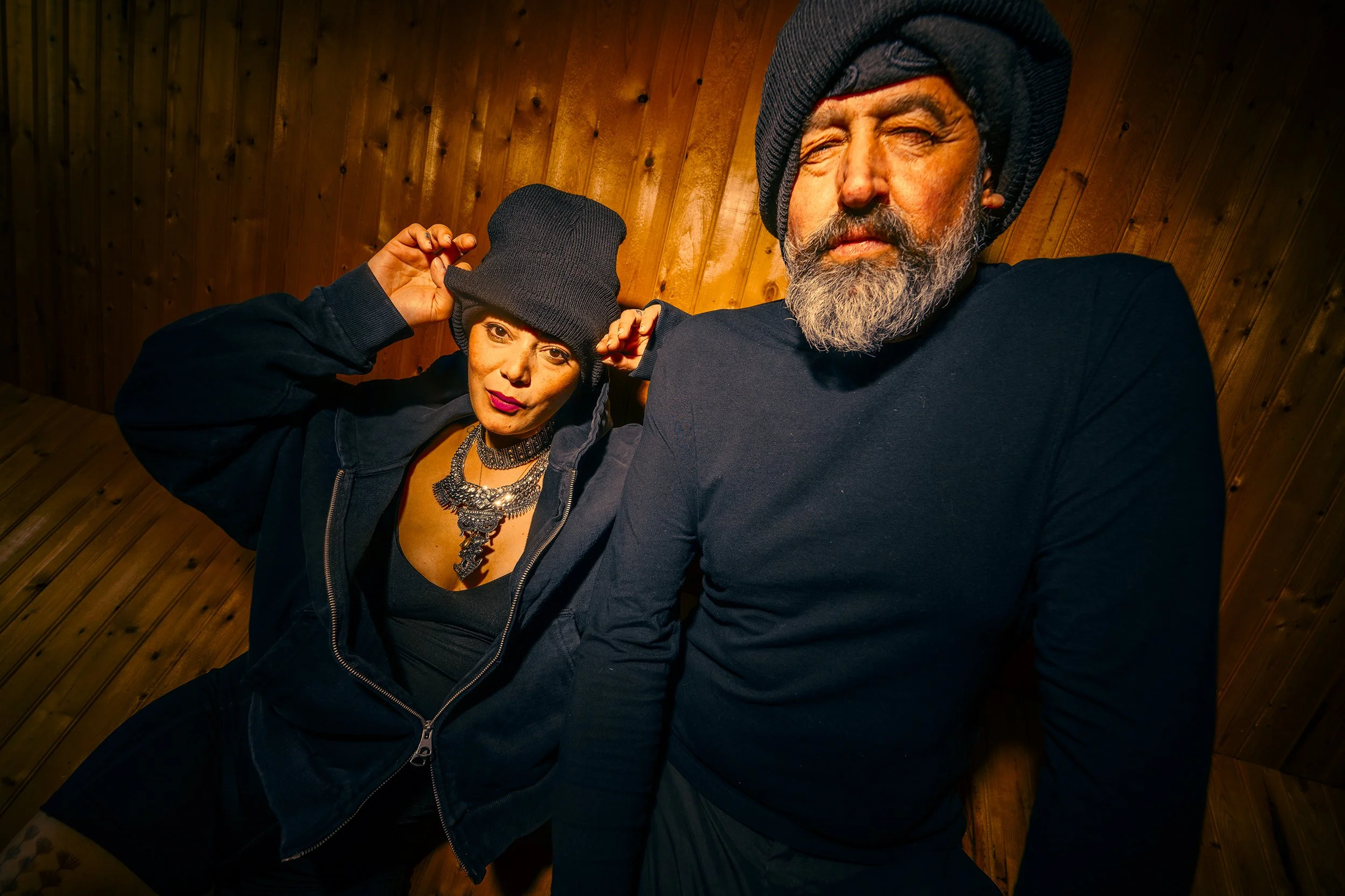 A woman and a man wearing black hoodies and black beanies, with the woman adjusting her beanie and the man with a gray beard and mustache, in front of a wooden wall.