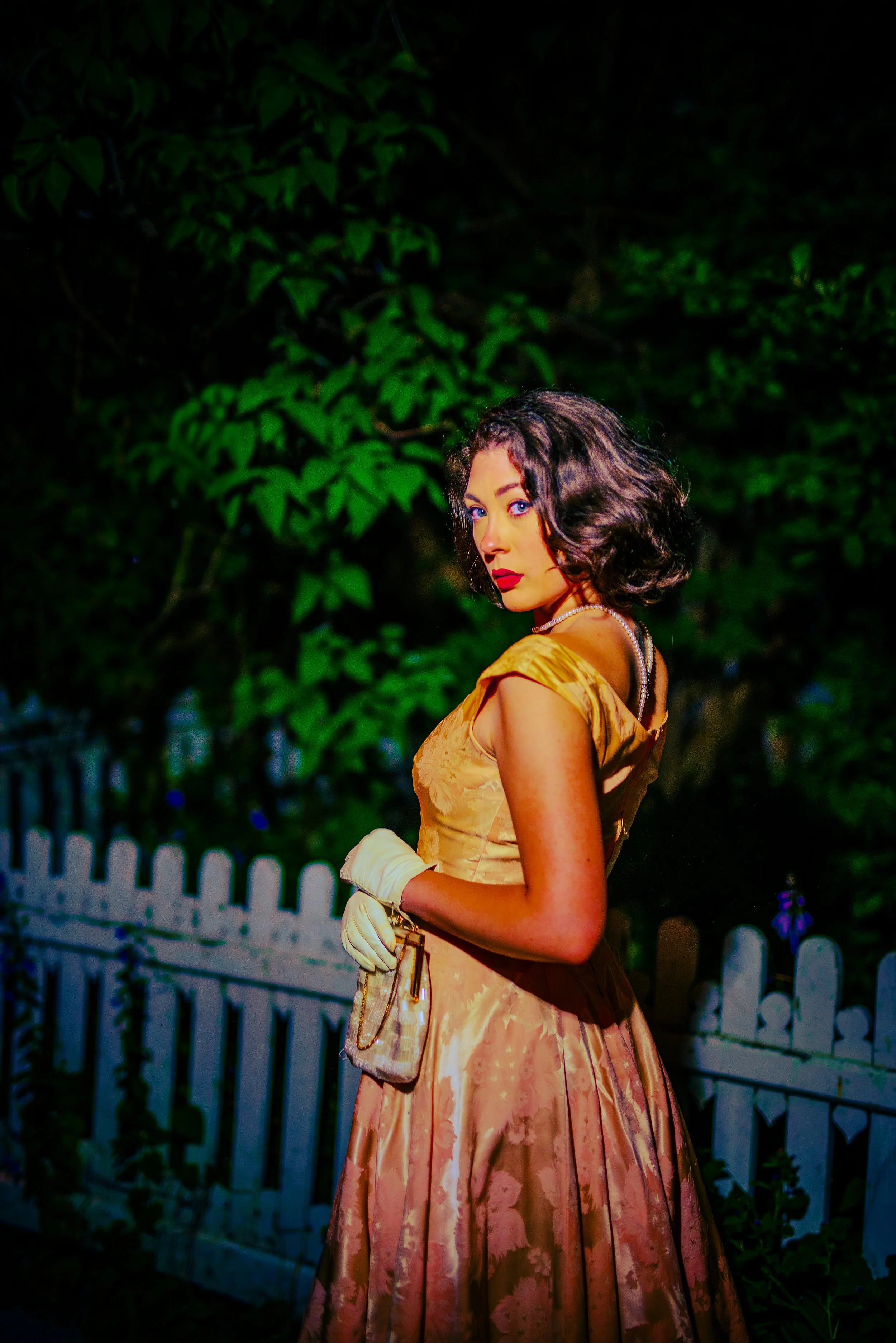 A woman in vintage 1950s style dress with floral pattern, pearl necklace, and vintage gloves, standing outdoors at night with green foliage and white picket fence in the background.