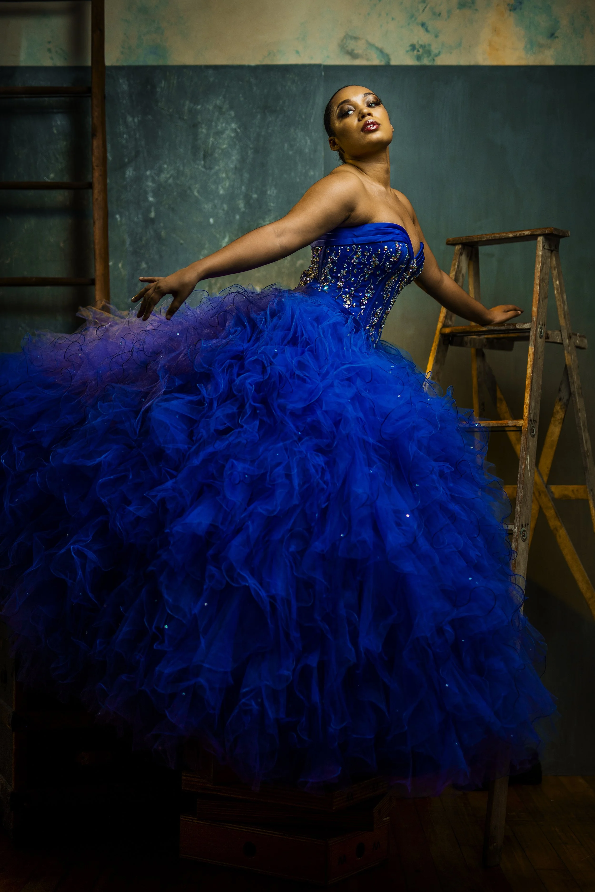 A woman in a blue strapless gown with a ruffled, voluminous skirt poses indoors near a wooden ladder and a wooden step stool.