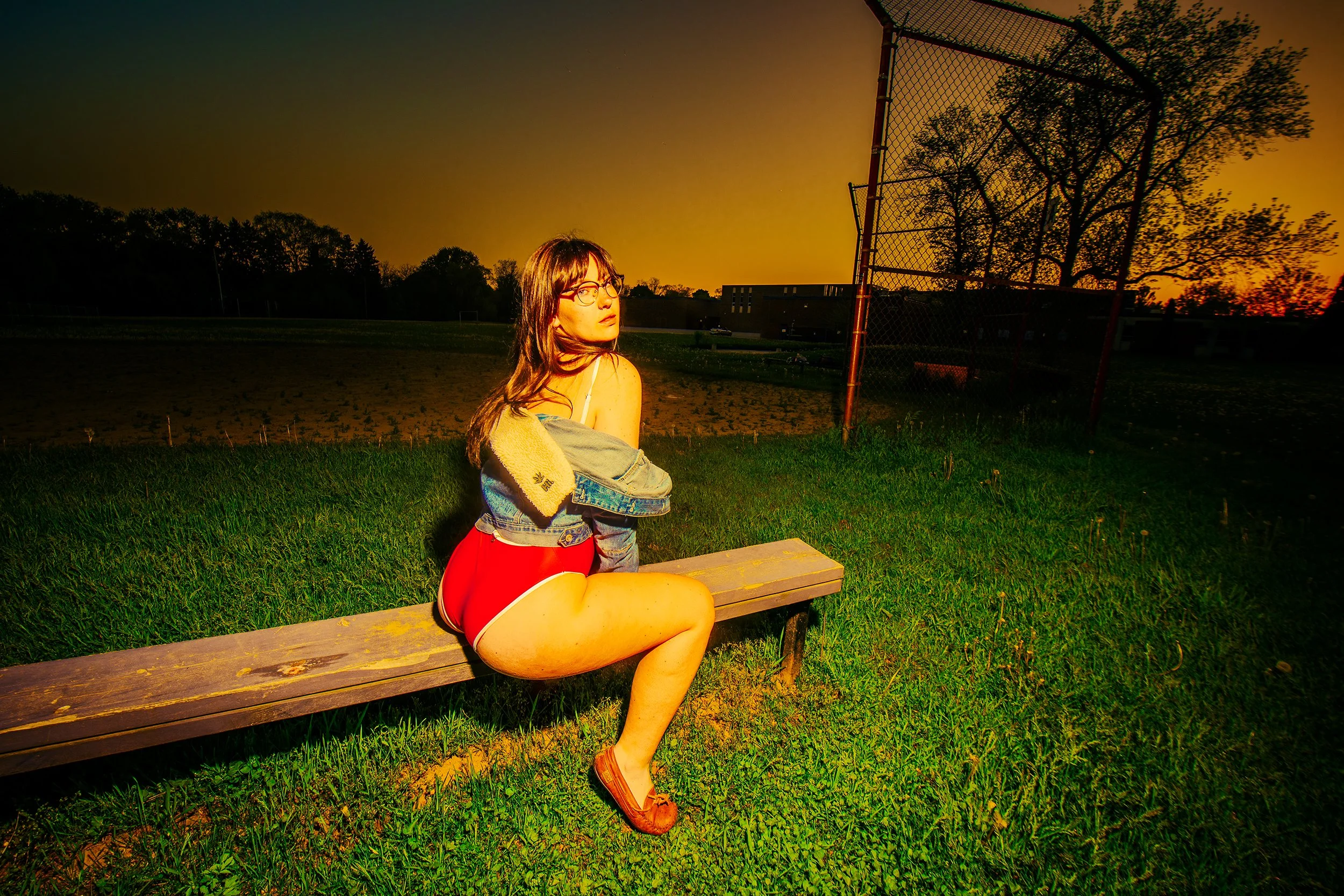 A young woman with glasses and long brown hair, sitting on a wooden bench at sunset, wearing a denim jacket and red shorts, with a towel draped over her shoulders, in a park with trees and a chain-link fence.