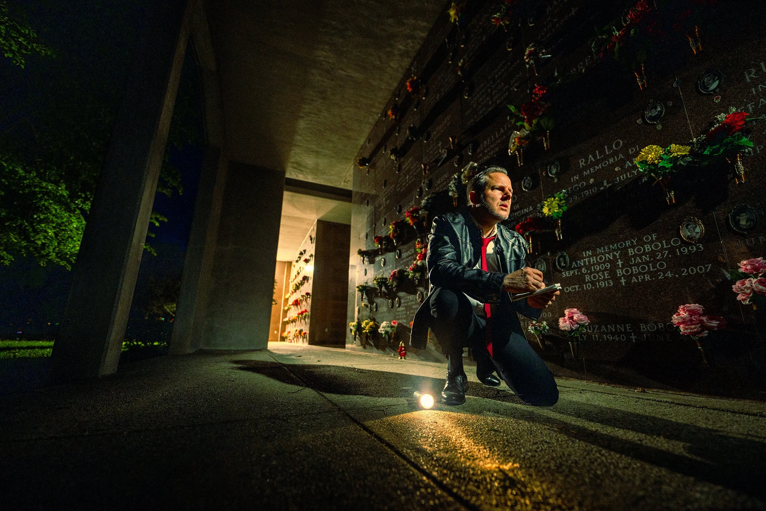 A man in a leather jacket kneels at night in front of a memorial wall, lighting a small candle. The wall is decorated with flowers and engraved names.