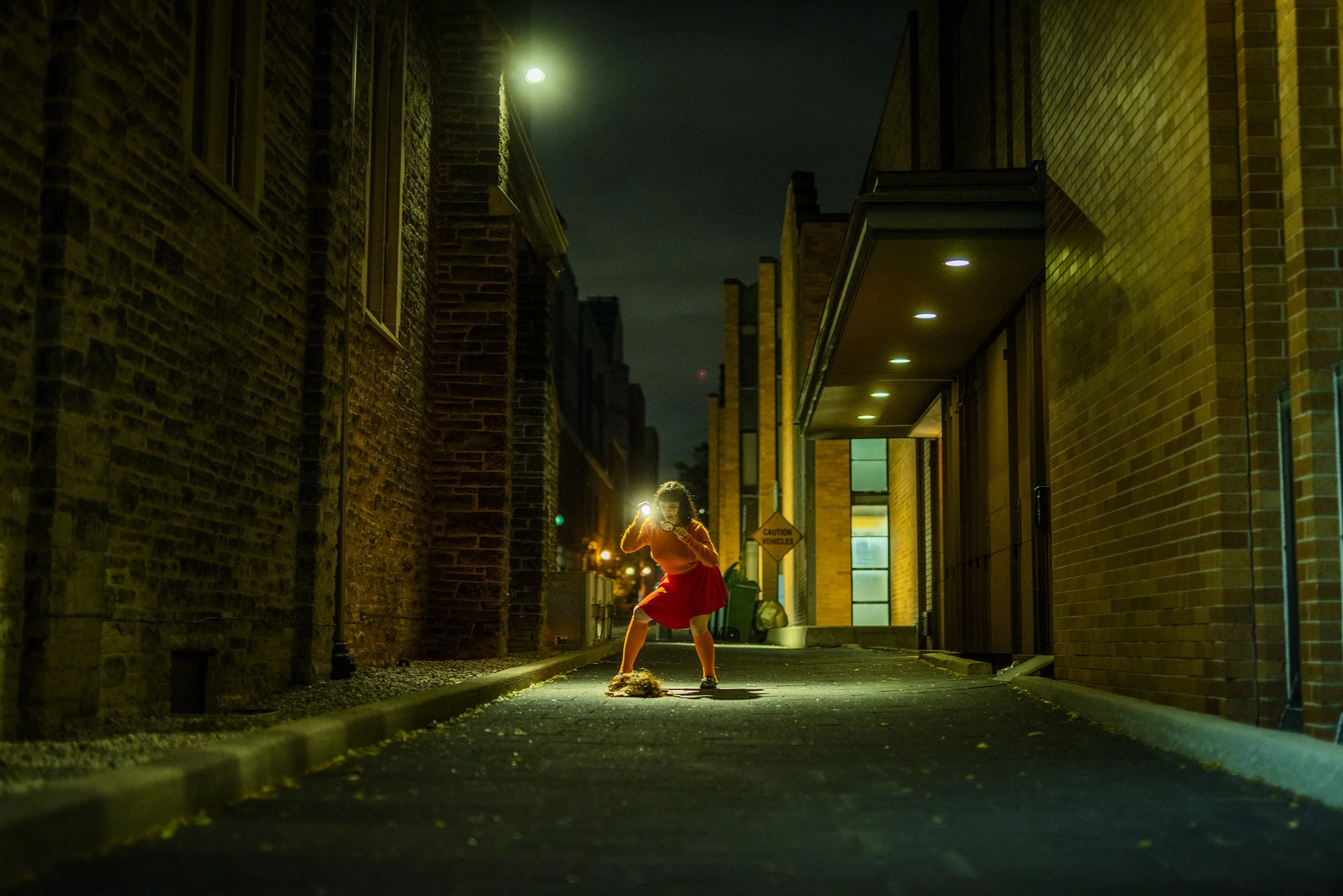 A woman in an orange top and red skirt is standing on a city street at night, illuminated by streetlights. She appears to be looking at her phone, with her hair covering her face. The scene is framed by tall brick buildings on either side and a build