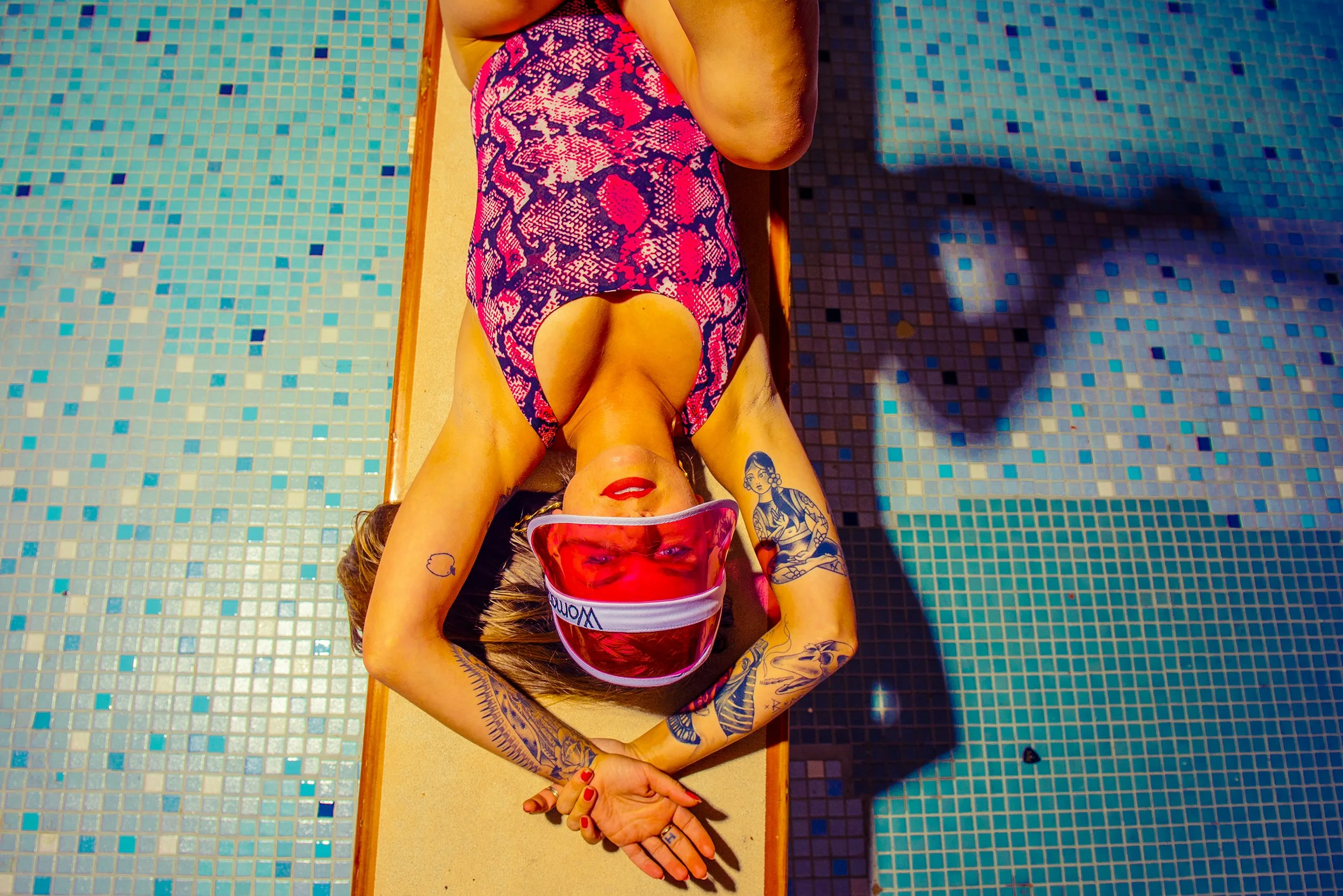 A woman lying on a poolside lounger with her upper body against the edge of the lounge chair, wearing a red and pink patterned swimsuit and a red visor. She has tattoos on her arms and is relaxing by a tiled swimming pool, with her eyes closed and he