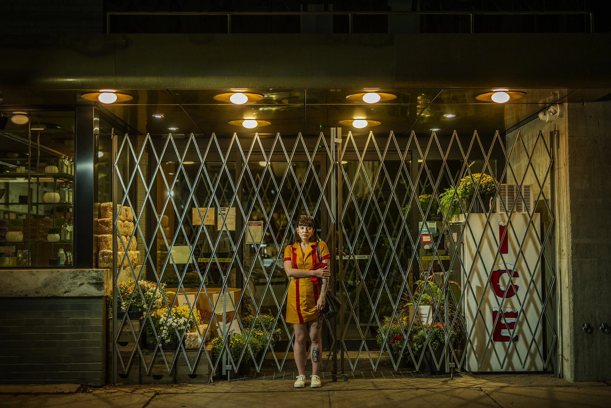 A woman in a yellow and red uniform standing outside a closed flower shop at night, holding a shopping bag and an umbrella.
